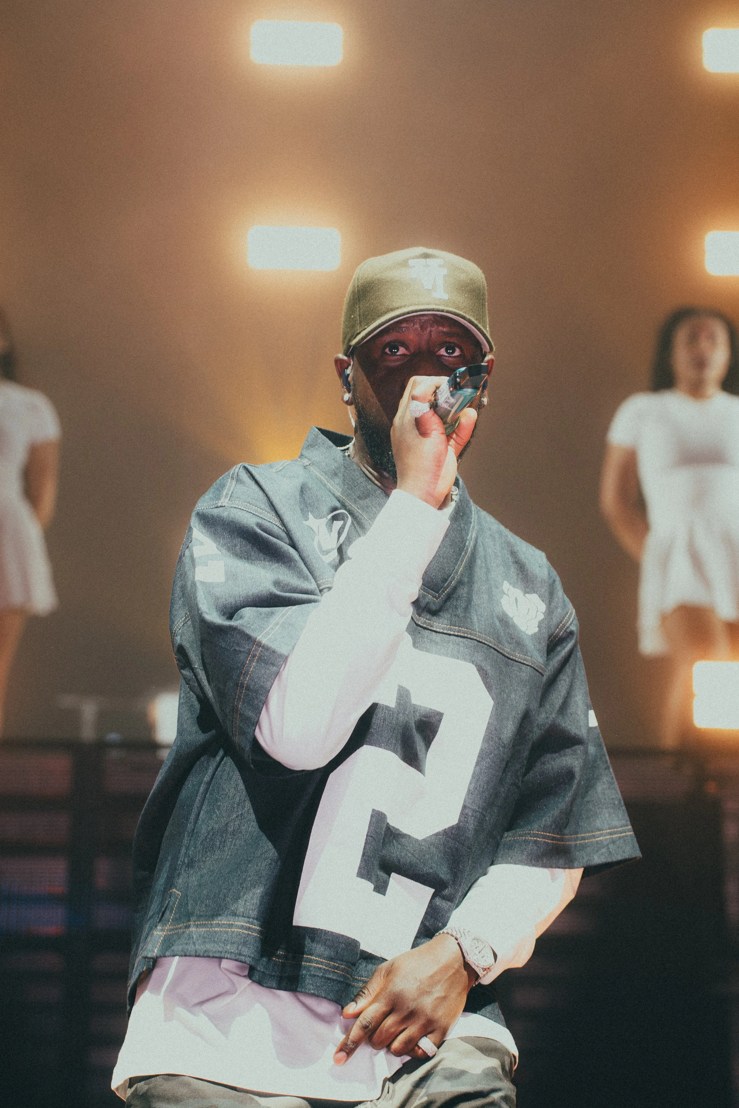 Un homme portant une casquette verte, un jean oversize avec le numéro 2, et tenant un microphone, en train de chanter ou parler sur scène, avec deux femmes en robe blanche floues en arrière-plan.