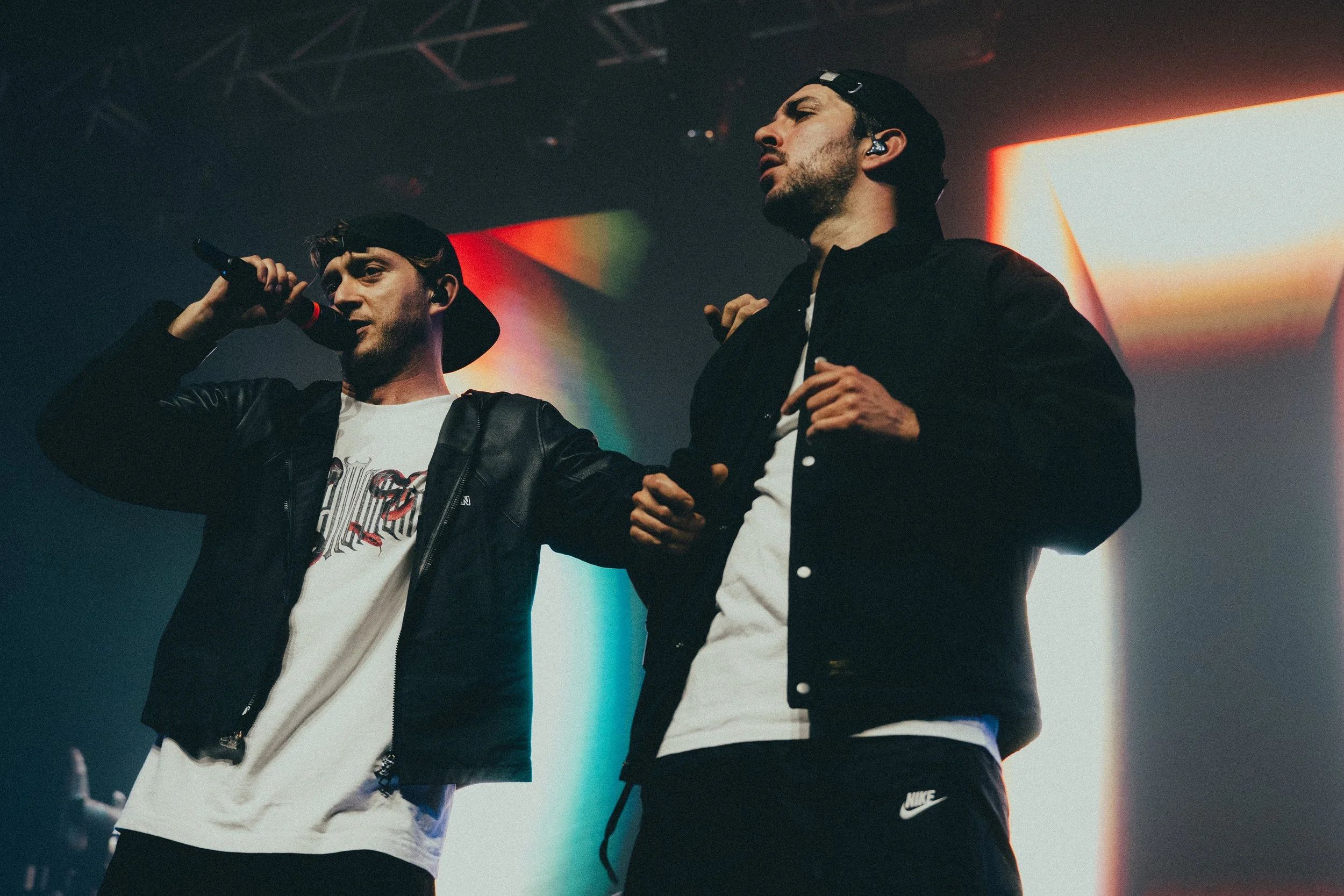 Deux jeunes hommes en vêtements décontractés chantent ou parlent sur scène lors d'une performance, avec un fond lumineux et coloré.