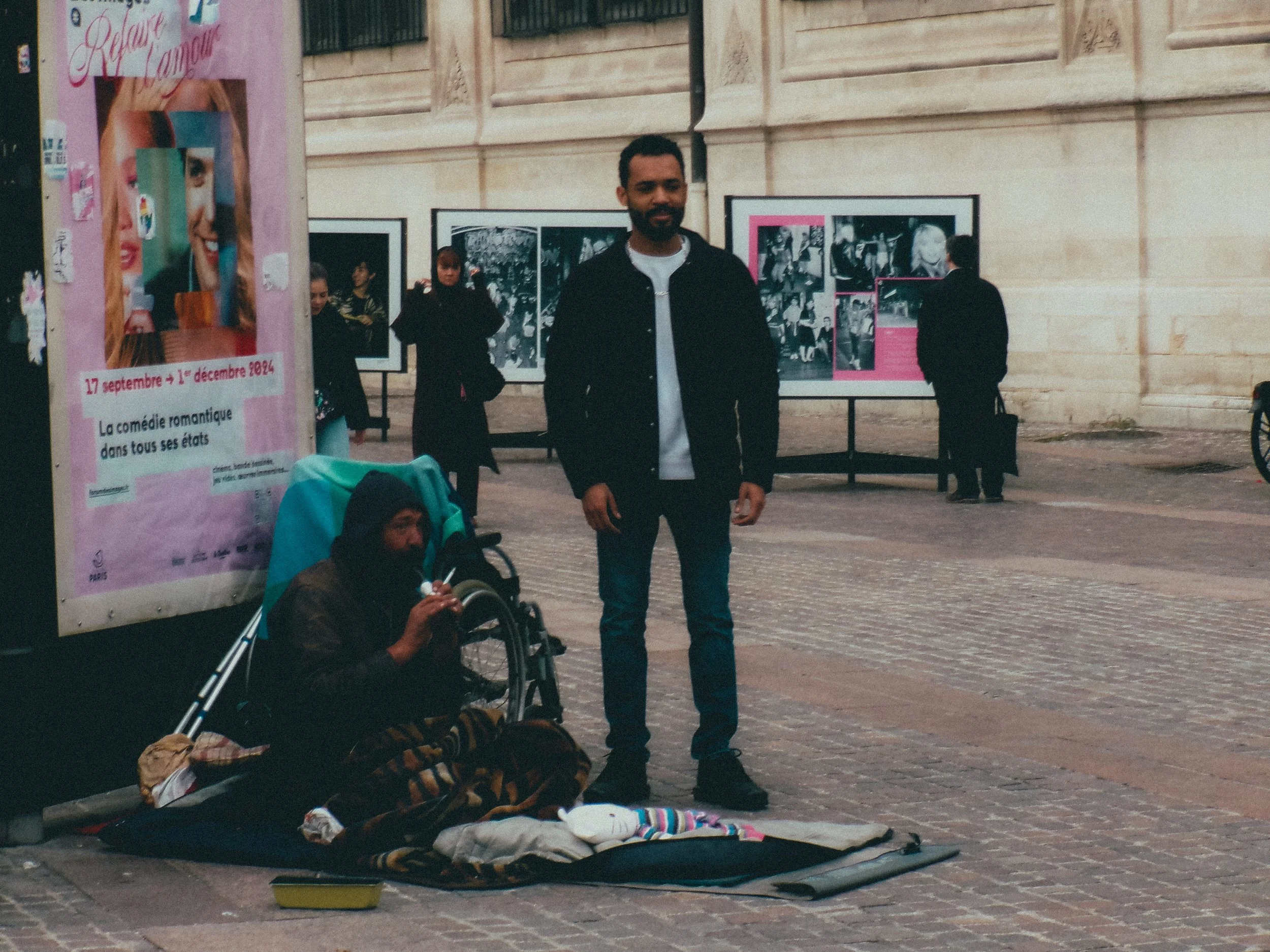 Un homme debout près d'un sans-abri qui est assis dans une rue pavée, avec des affiches d'une exposition photographique en arrière-plan.