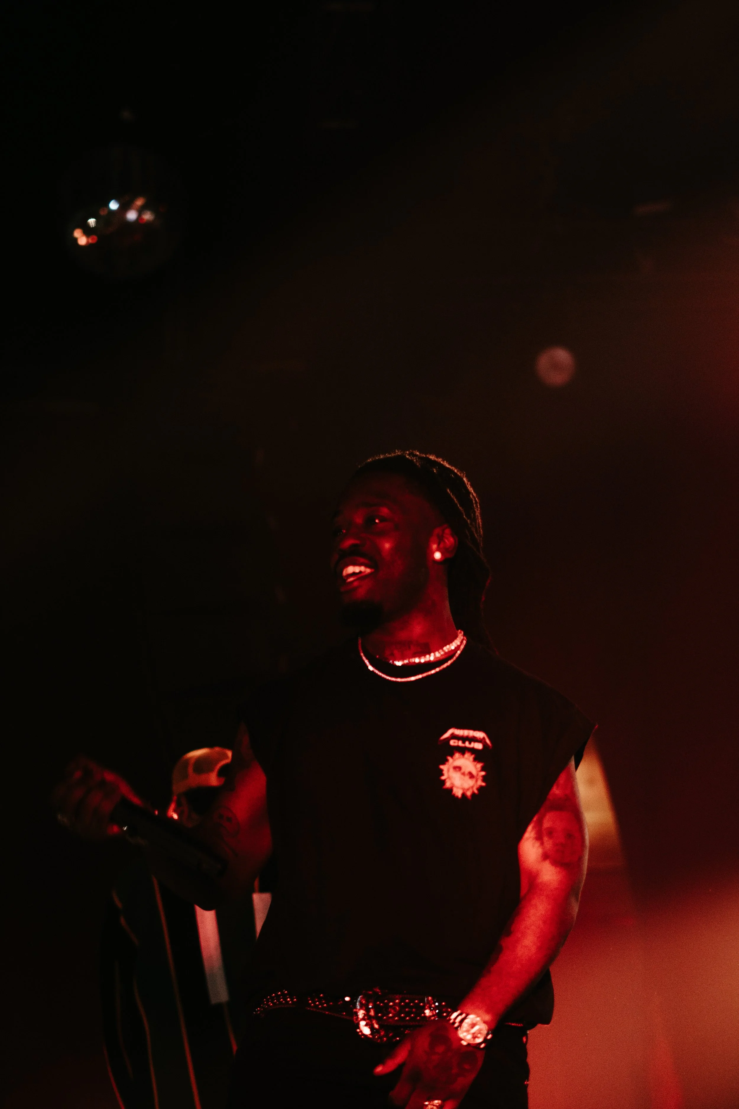 Homme noir avec dreadlocks, en noir, portant des bijoux, dans un environnement sombre avec des lumières rouges, tenant un micro.
