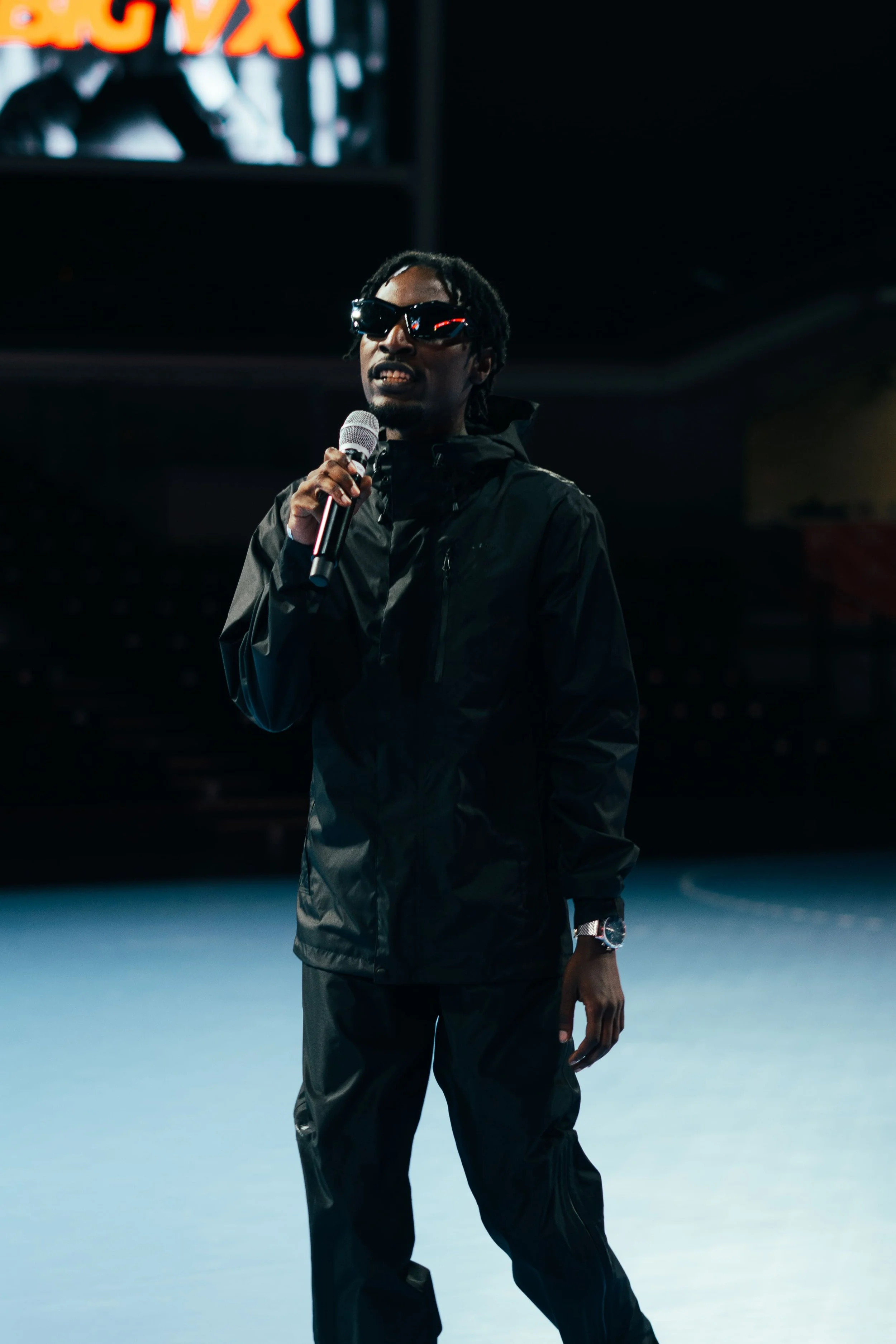 Un homme portant des vêtements noirs, des lunettes de soleil, tenant un micro, se tenant sur une scène à l'intérieur.