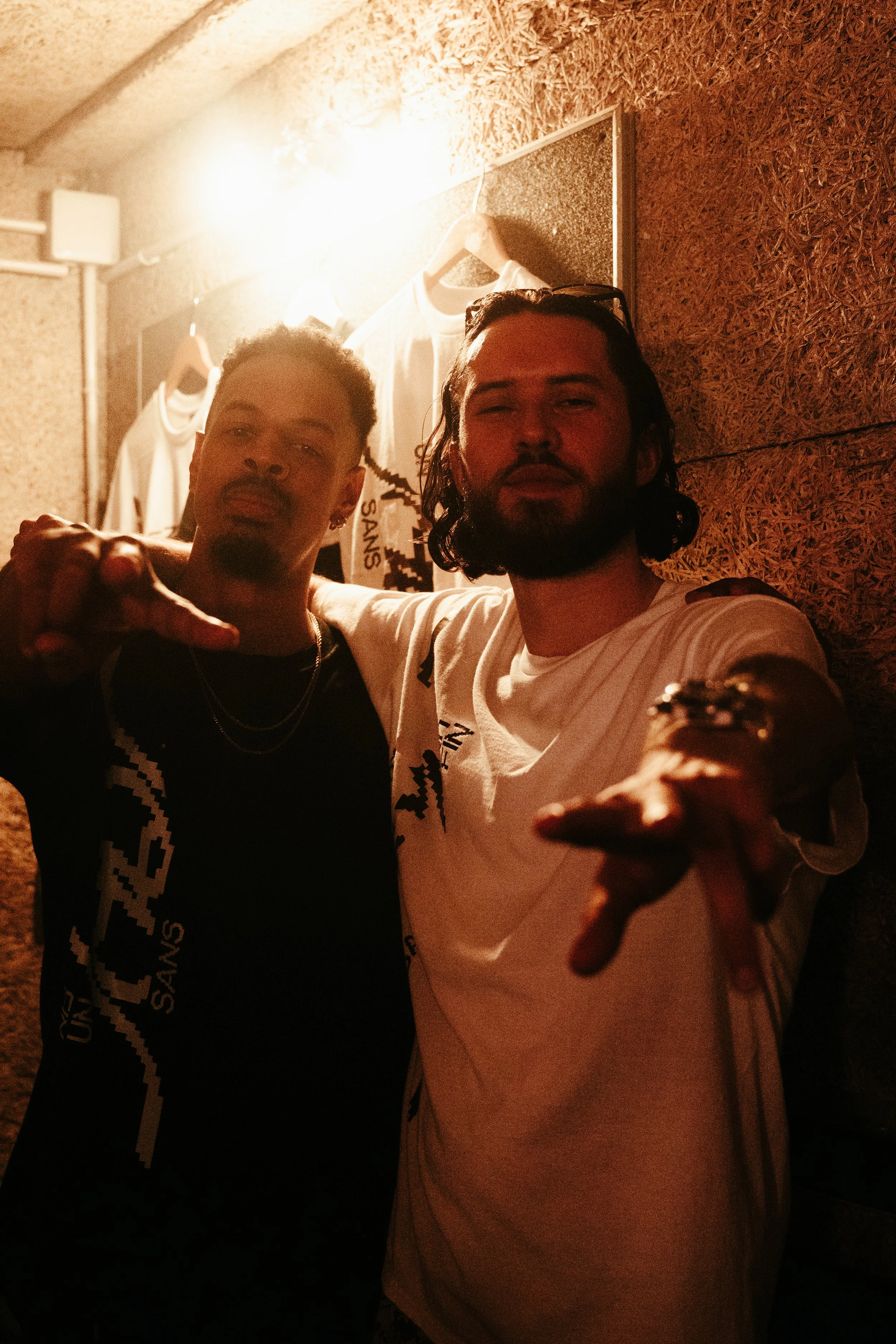 Deux hommes posant ensemble, l'un en t-shirt blanc et l'autre en t-shirt noir, dans un endroit avec un mur en bois et des vêtements suspendus en arrière-plan.