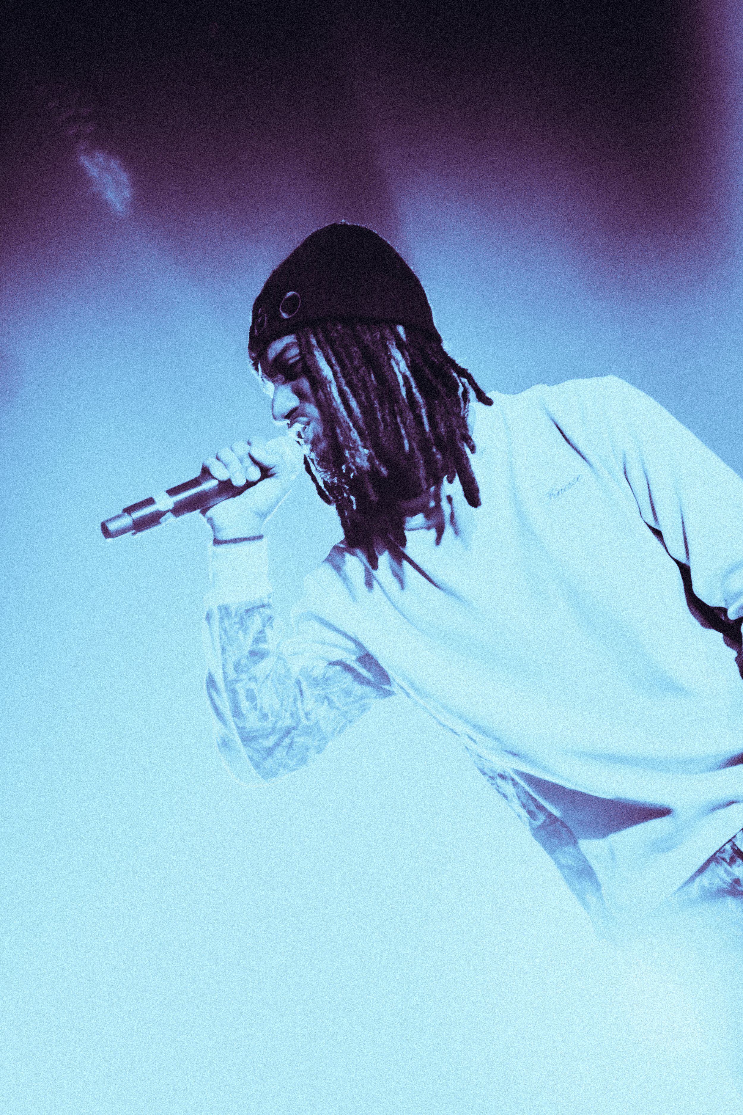 Un homme avec des dreadlocks porte un bonnet noir et tient un micro, en train de chanter ou de performer, dans une ambiance sombre avec un éclairage bleu.