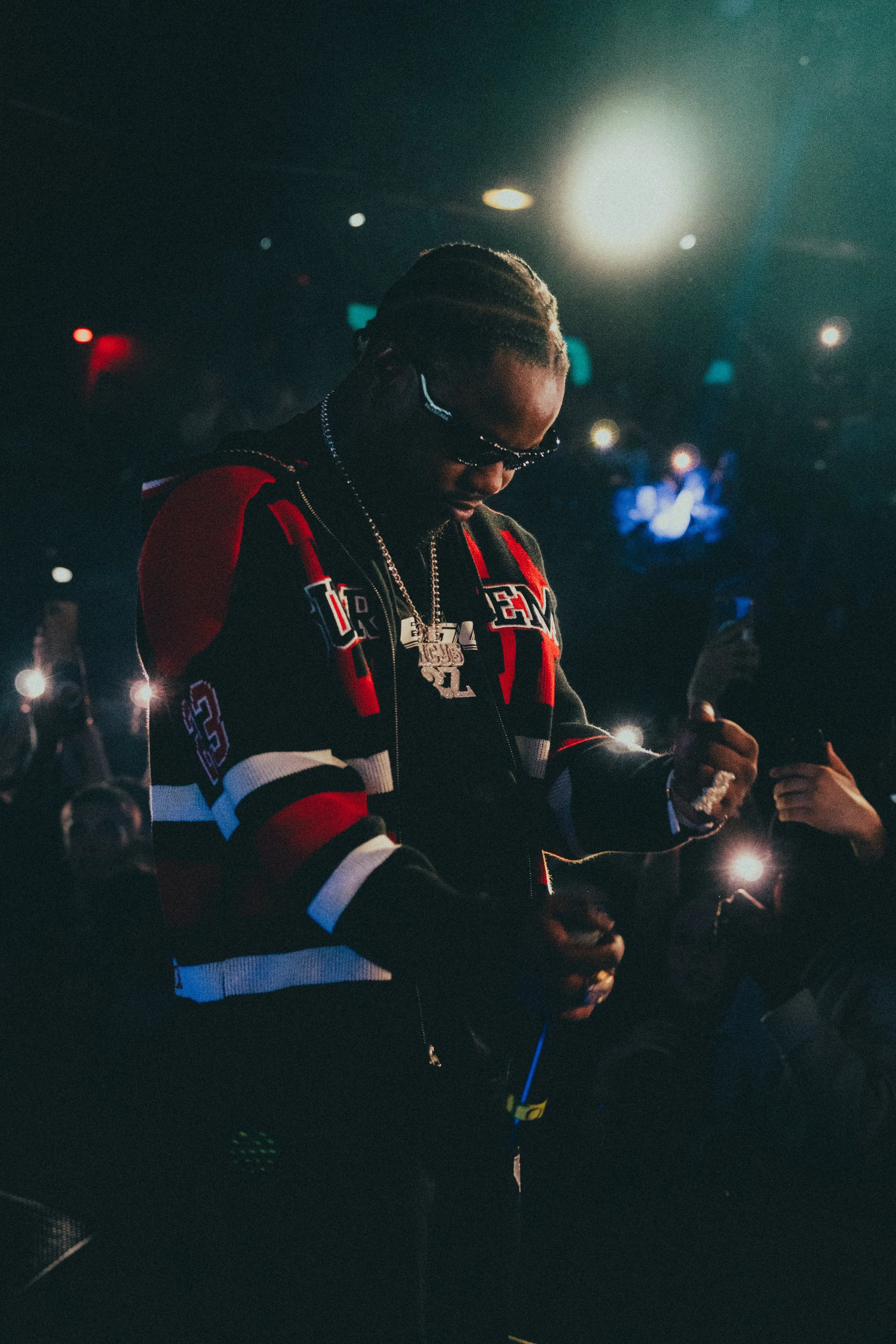 Un homme portant des vêtements de sport, avec des bijoux, dans un environnement sombre et brillant, probablement lors d'un événement ou concert.