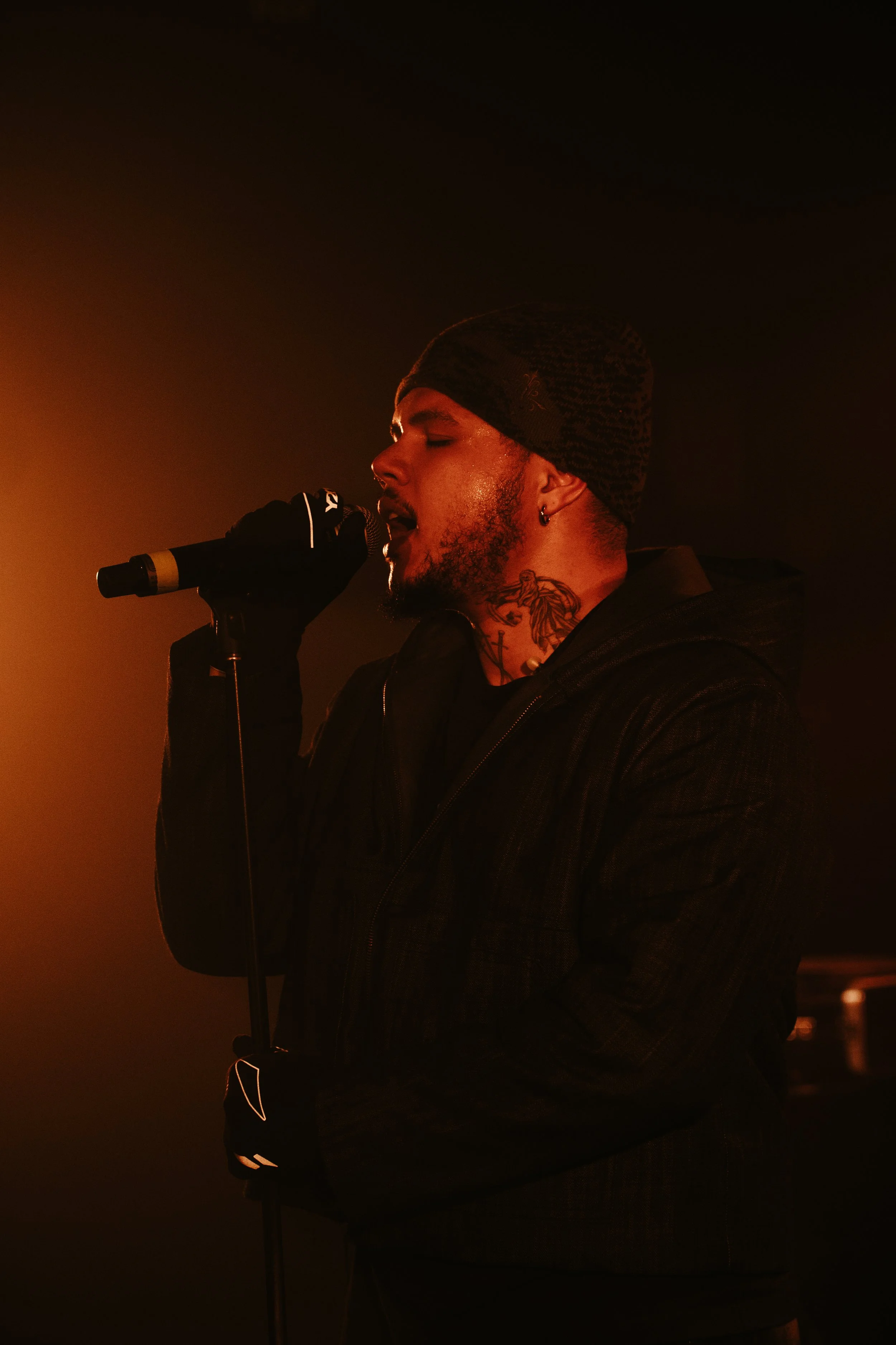 Un homme chante dans un micro, portant un bonnet, un gilet noir et des tatouages visibles sur le cou, dans une ambiance sombre et chaude.