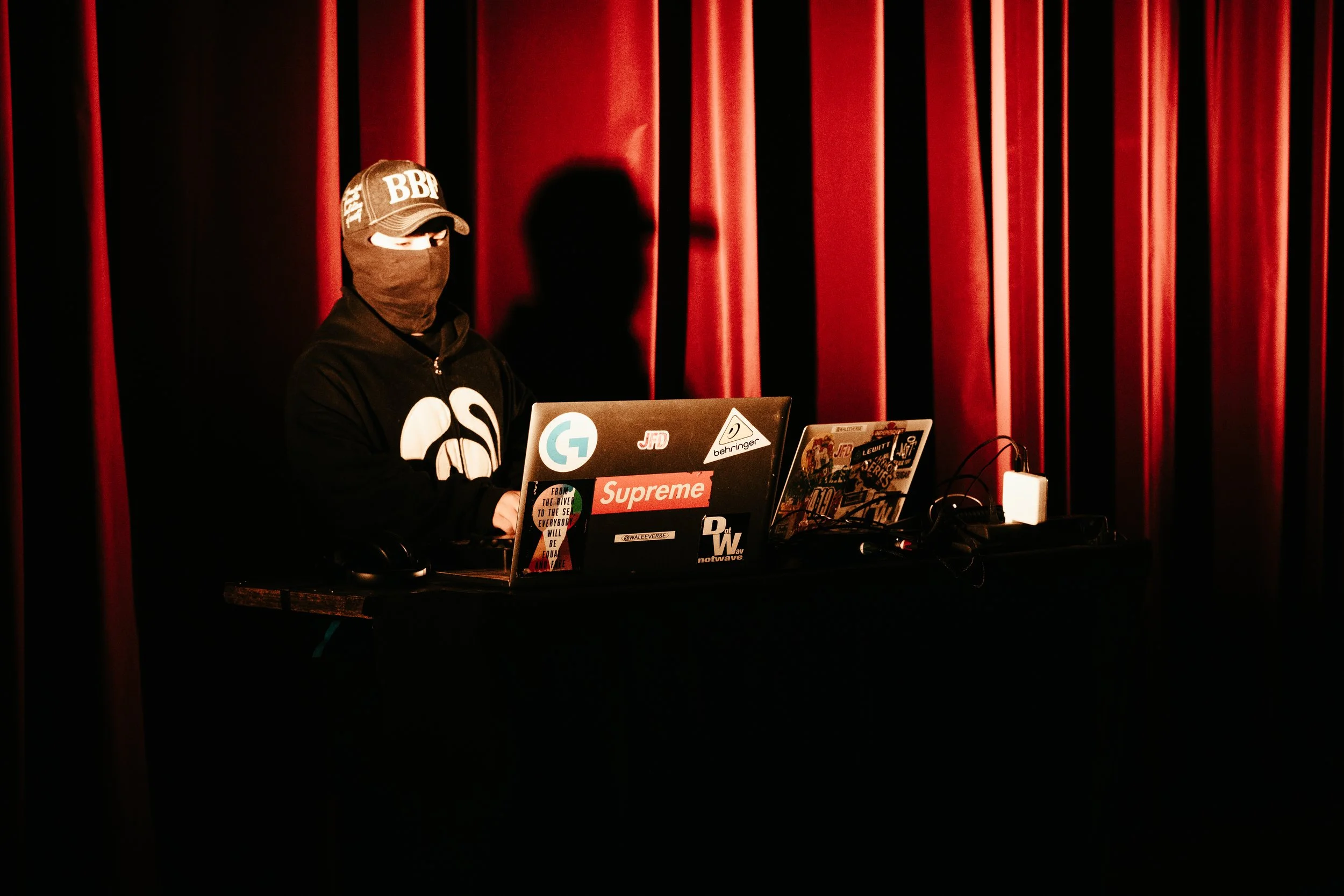 Un individu masqué portant une casquette et un sweat-shirt noir, travaillant sur un ordinateur portable décoré de nombreux autocollants, dans un environnement sombre avec rideaux rouges en arrière-plan.