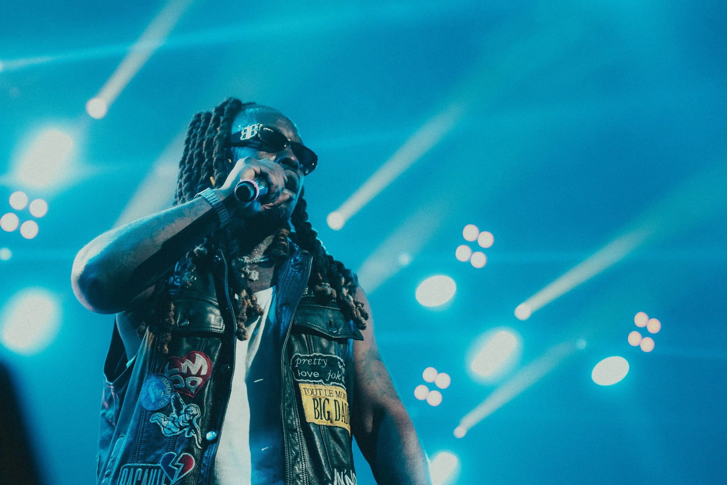 Un artiste masculin avec des dreadlocks, portant des lunettes de soleil et une veste en cuir décorée de patches, chante avec un microphone sur scène avec des lumières de concert en arrière-plan.