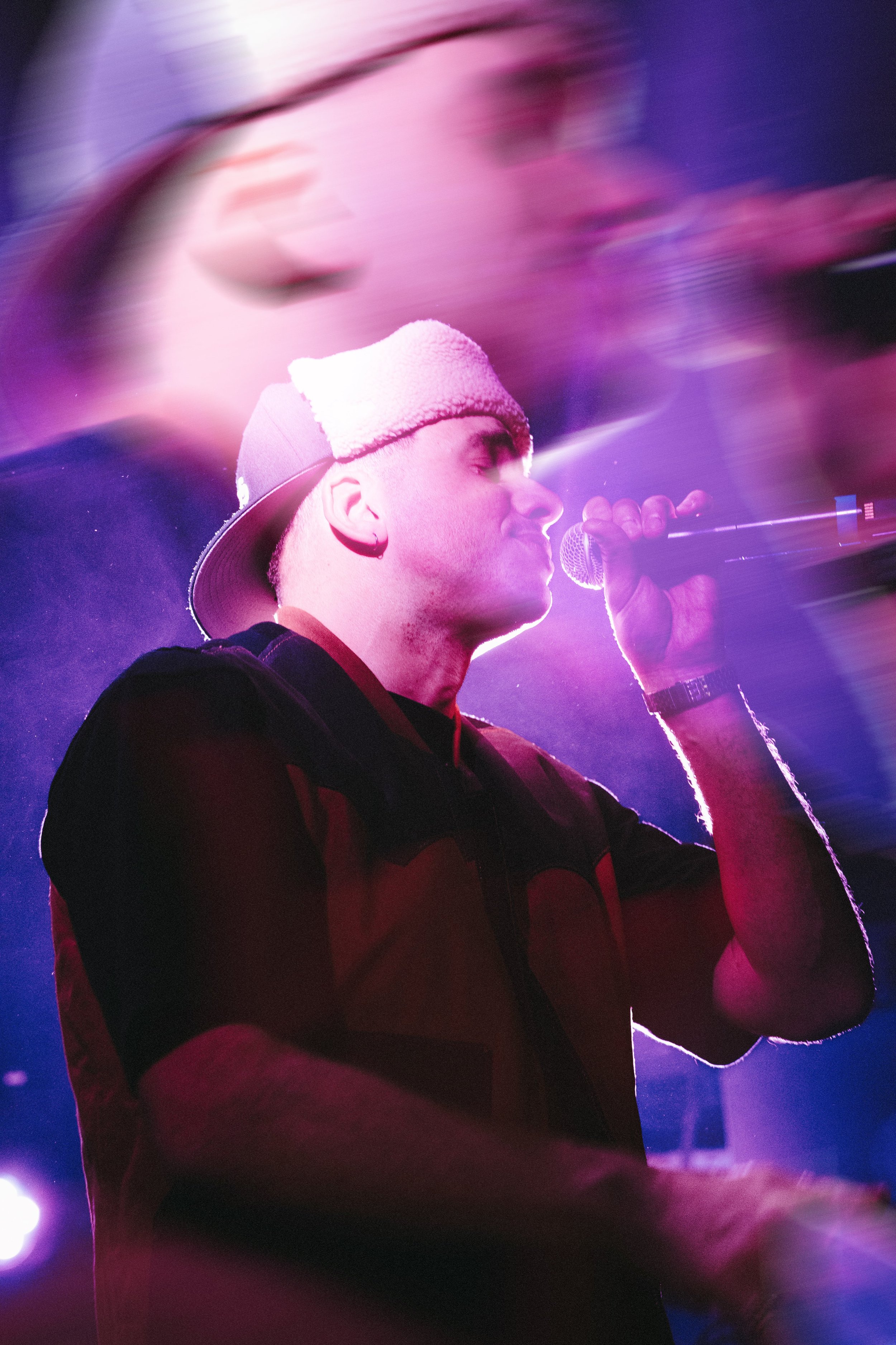 Un homme portant une casquette de baseball blanche, chantant ou parlant dans un microphone, avec un éclairage violet et rose sur la scène, et un effet de mouvement flou autour.