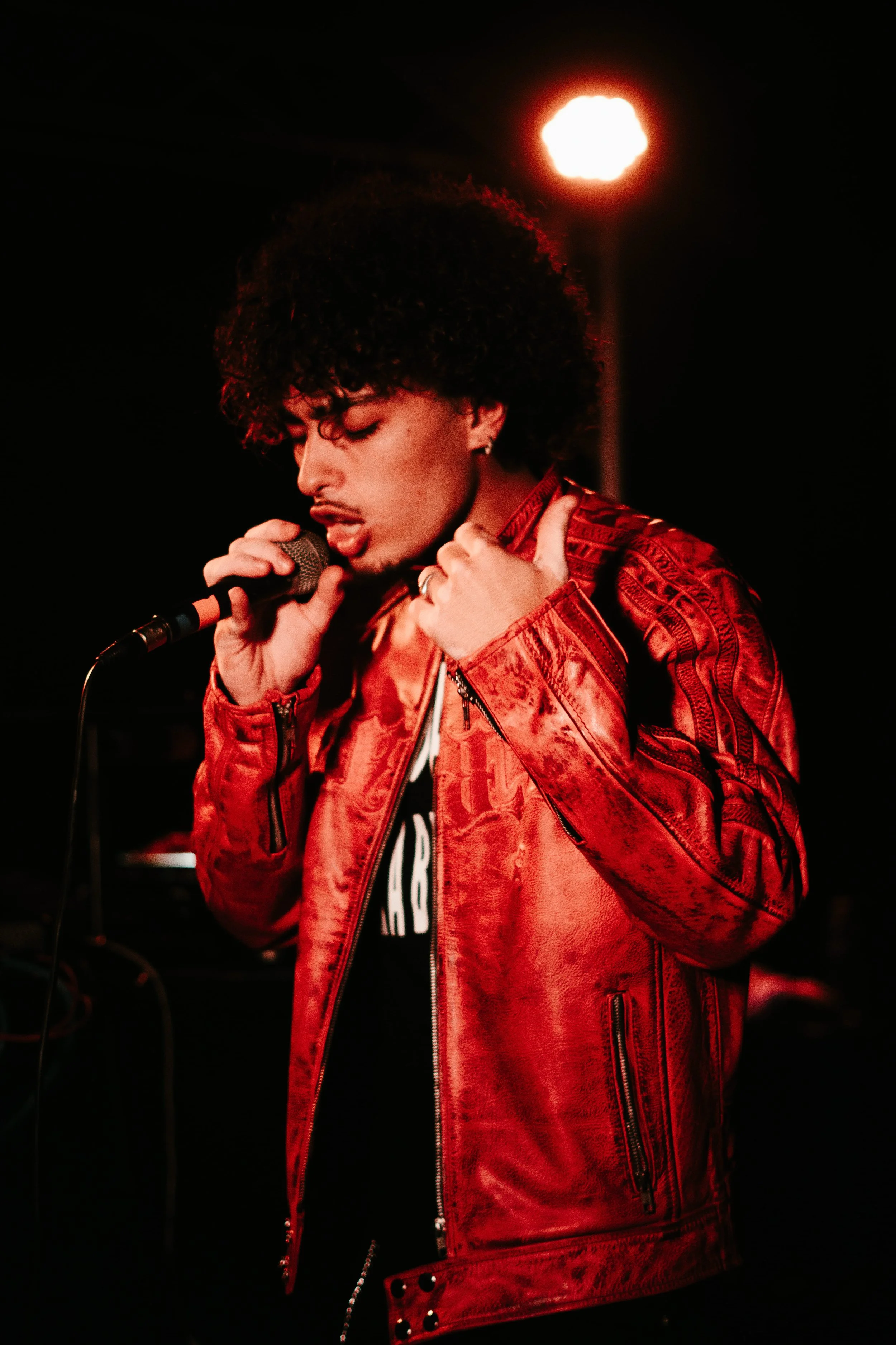 Un homme chante dans un micro, portant une veste rouge en cuir, avec un éclairage chaud en arrière-plan.