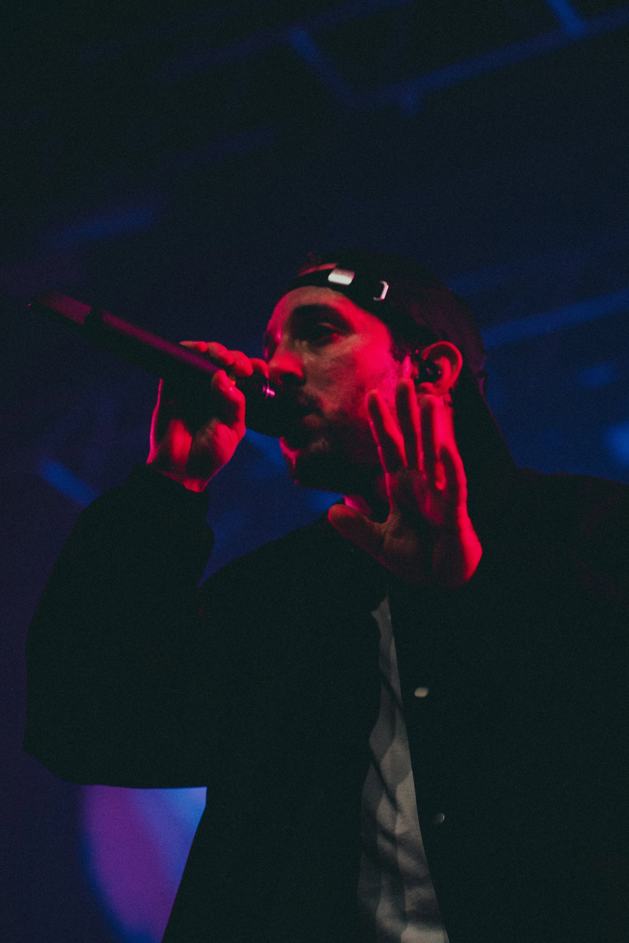 Un performer chante dans un micro sur scène, avec un éclairage rouge et bleu, portant une casquette noire rabattue vers l'avant.