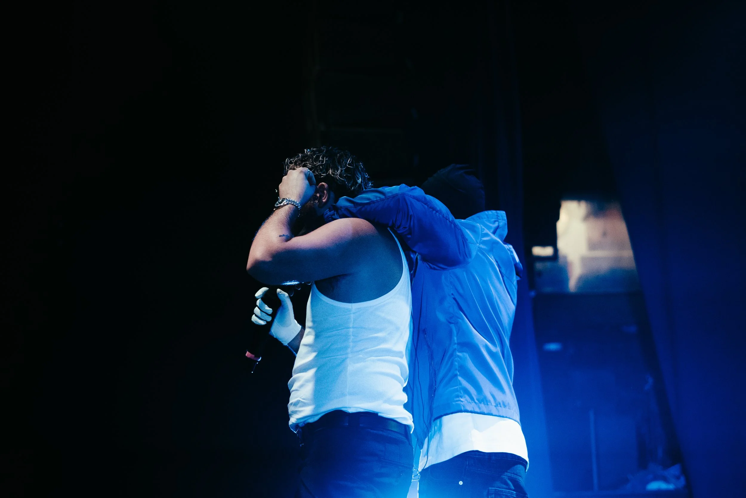 Deux personnes sur scène, dont une avec un micro, en tenue décontractée, dans une ambiance sombre avec un éclairage bleu.