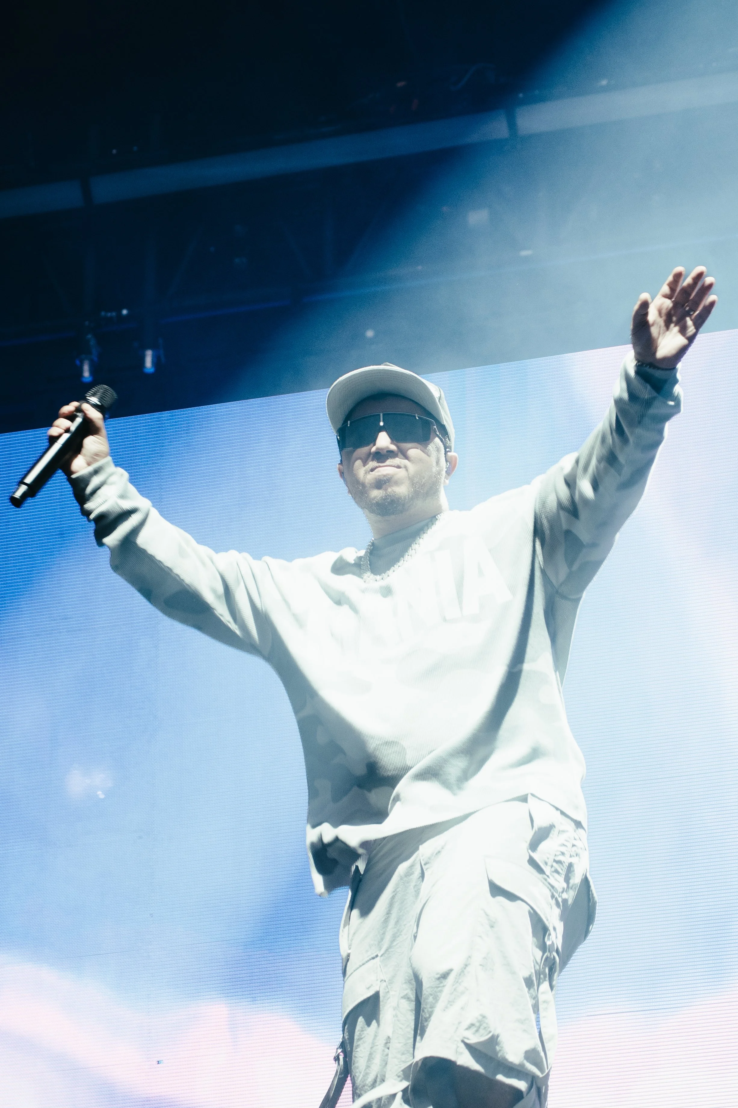 Un homme portant des vêtements décontractés, des lunettes de soleil et une casquette, tenant un microphone, sur scène lors d'un concert, avec un grand écran en arrière-plan.