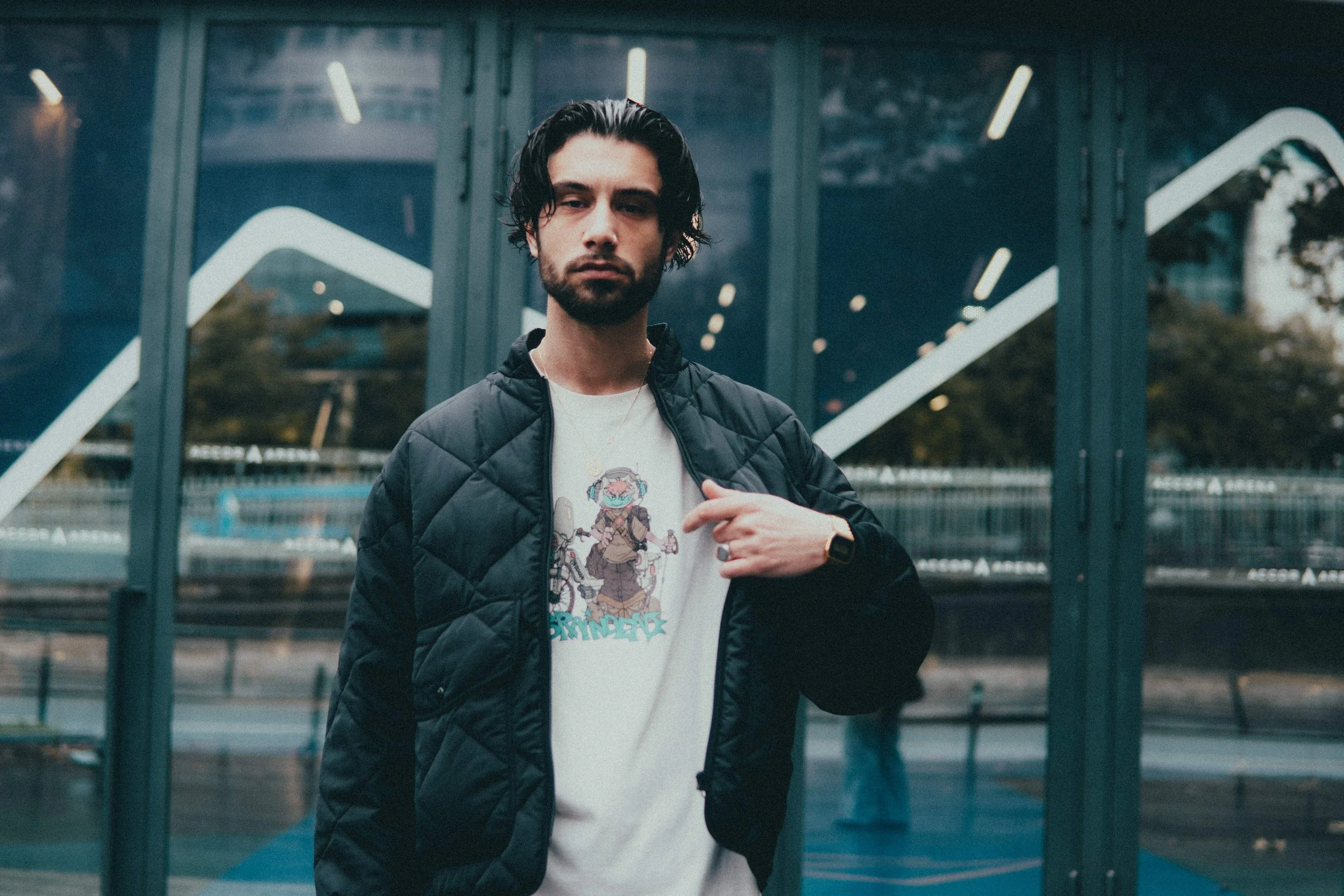 Un jeune homme avec des cheveux noirs et une barbe porte une veste noire et un t-shirt avec un dessin coloré. Il se tient devant une structure en verre dans une zone urbaine.