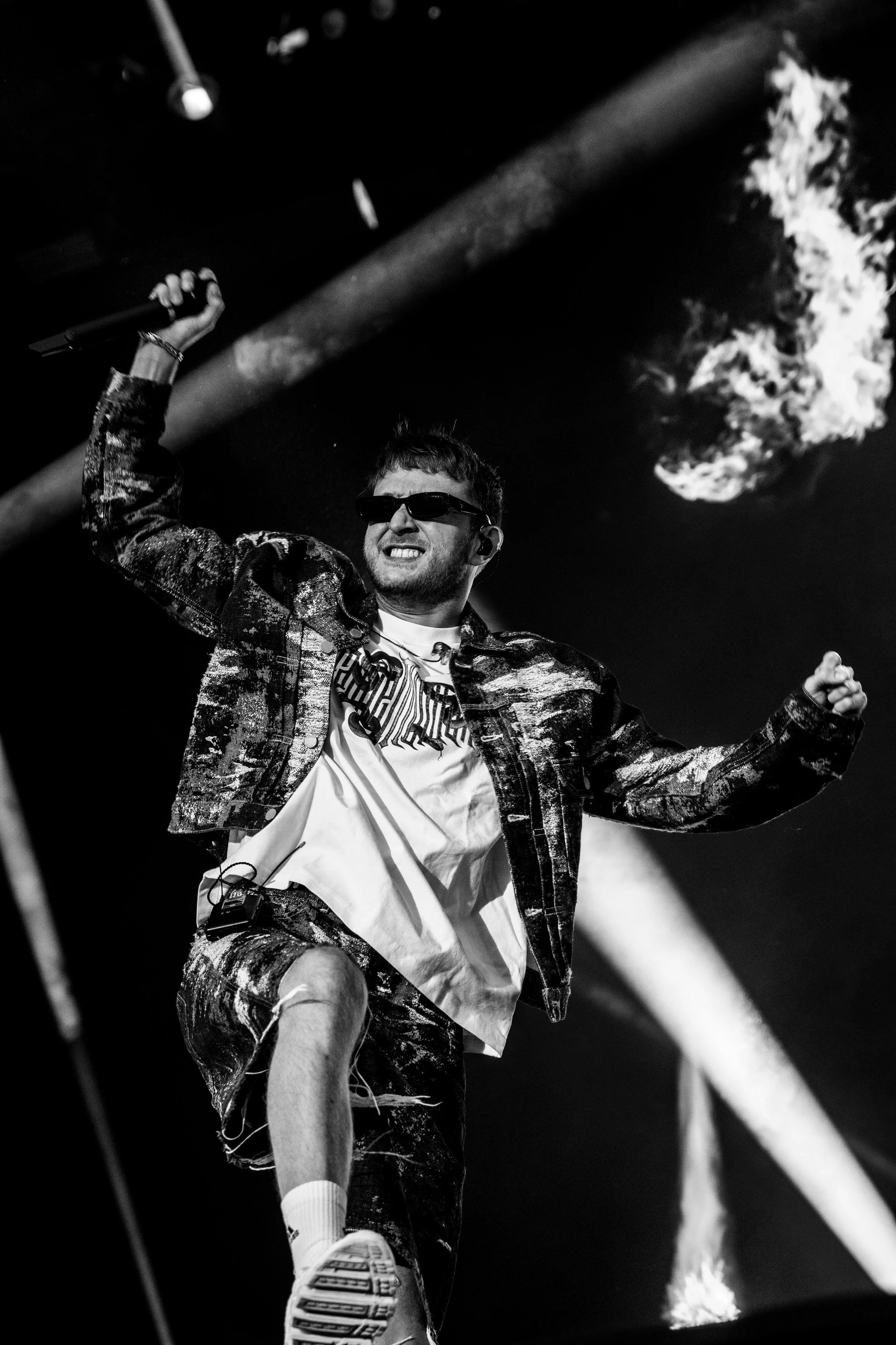 Un homme portant des lunettes de soleil, un pantalon déchiré, une chemise blanche et une veste à motifs, tenant un microphone en l'air lors d'une performance sur scène. La scène est éclairée par des projecteurs, avec des effets de lumière et un feu e