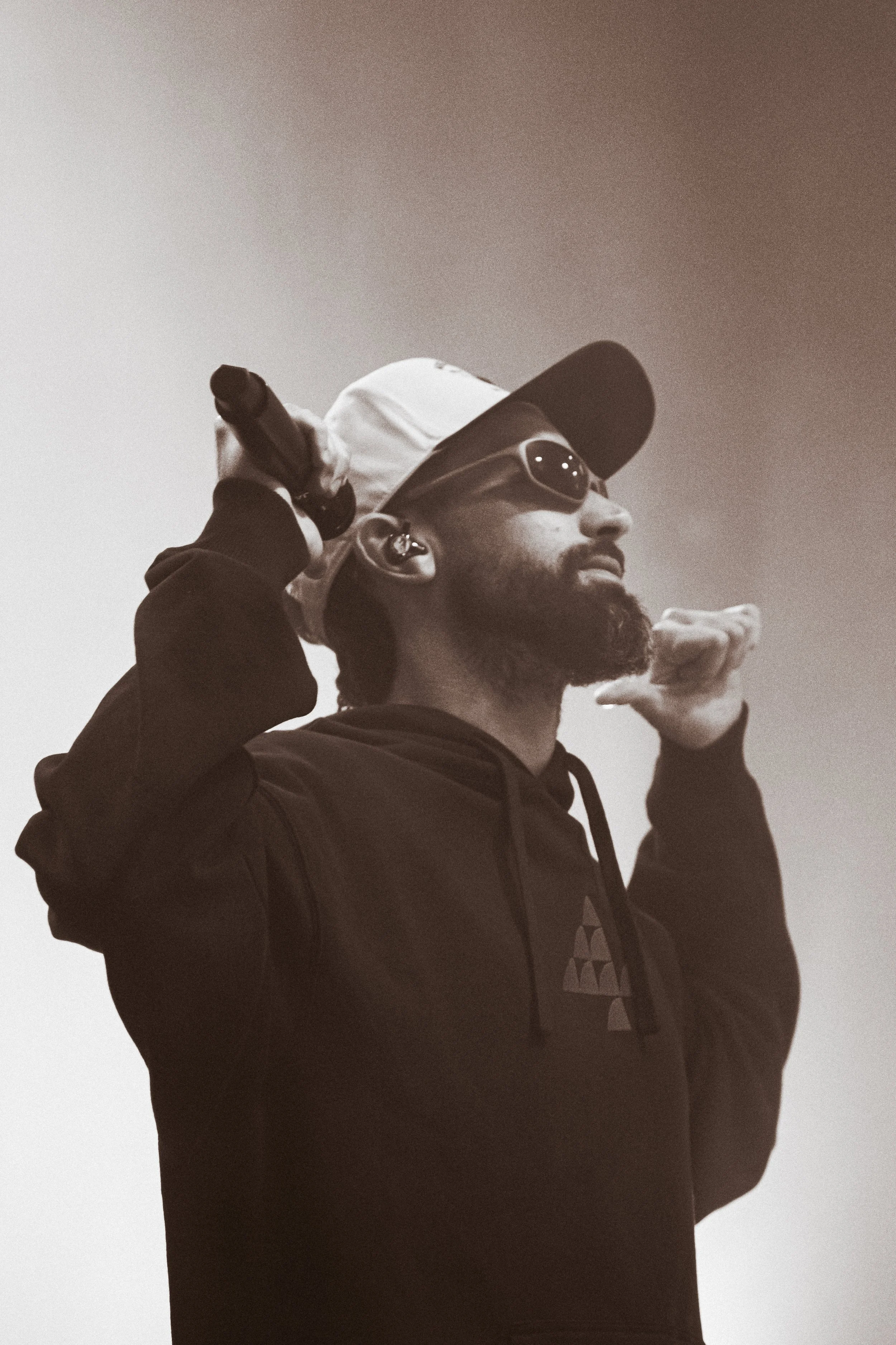 Un homme avec une barbe portant une casquette, des lunettes de soleil, un écouteur dans l'oreille, et un sweat à capuche, tenant un micro dans la main, en train de parler ou de chanter.