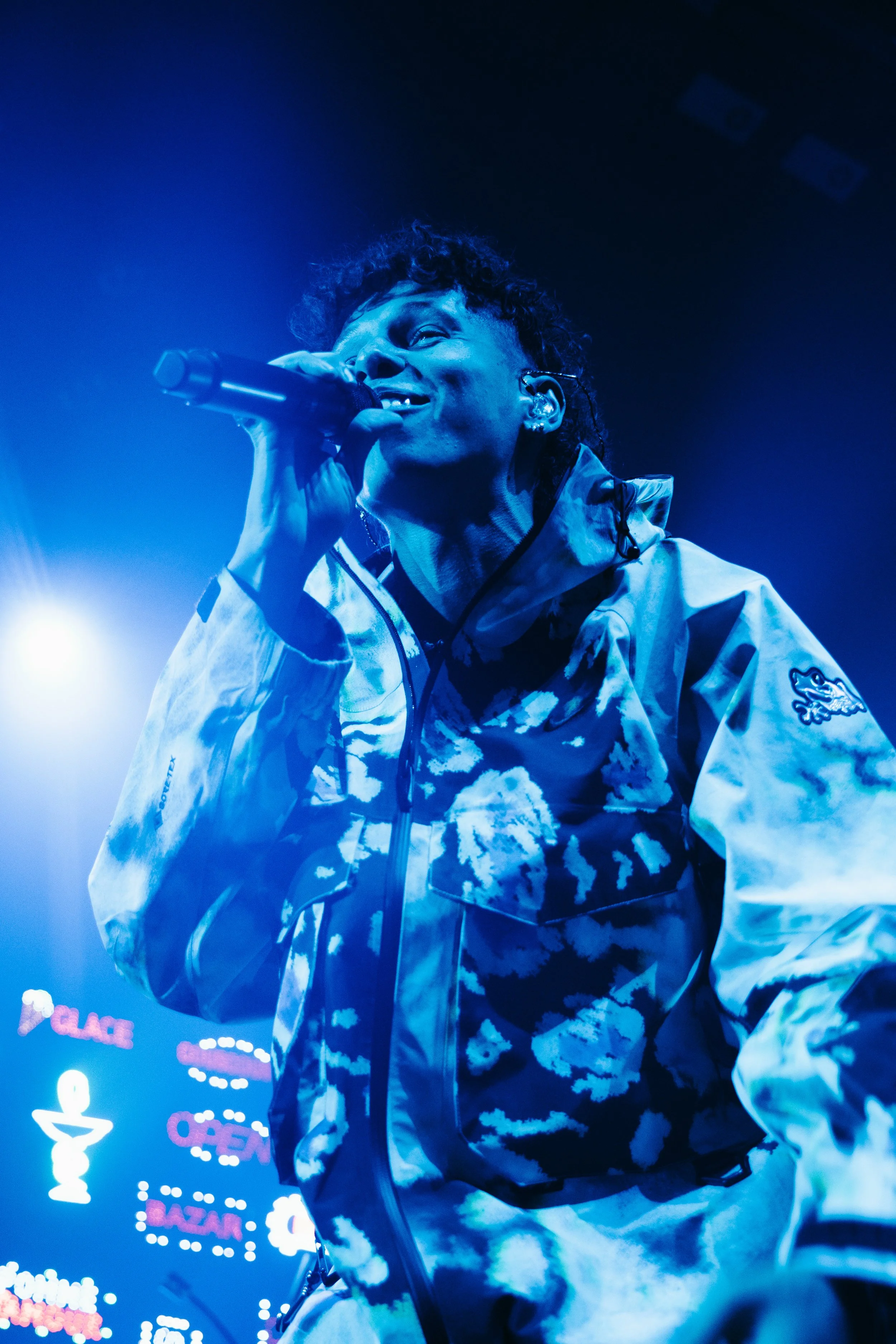 Un homme chante avec un microphone lors d'un concert, portant une veste à motif nuageux, sous un éclairage bleu.