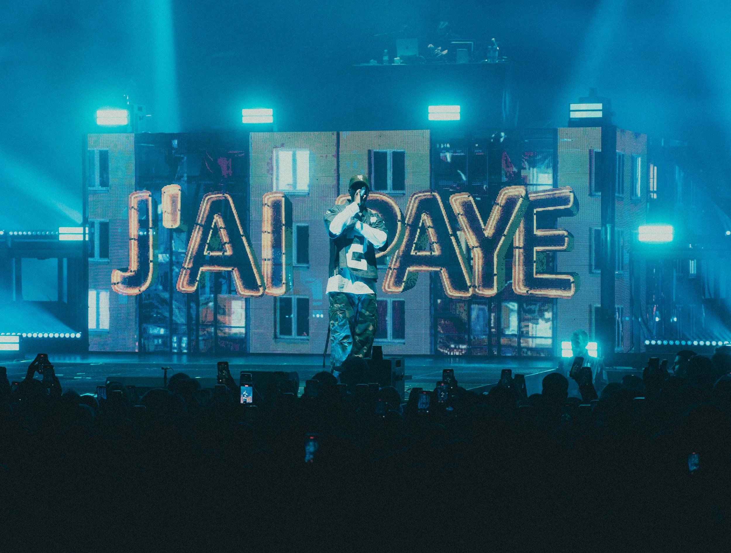 Un artiste en performance sur scène devant un grand écran lumineux avec le nom JAILAYE, dans une ambiance de concert avec des lumières bleues et un public pris en photo avec des téléphones.
