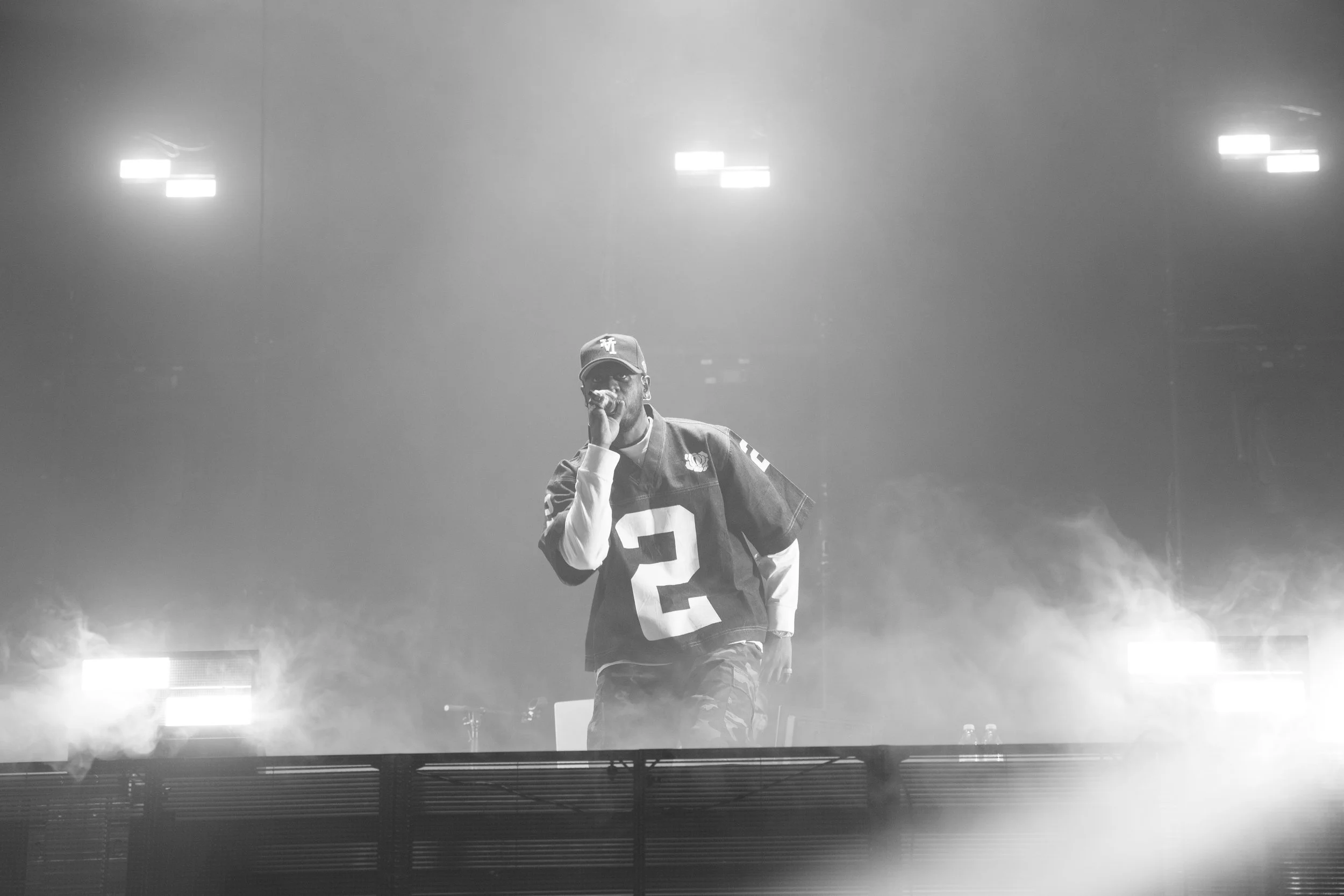 Homme en sweat à capuche et casquette, chantant ou parlant dans un micro, dans une ambiance de concert ou de scène avec de la fumée et des lumières.