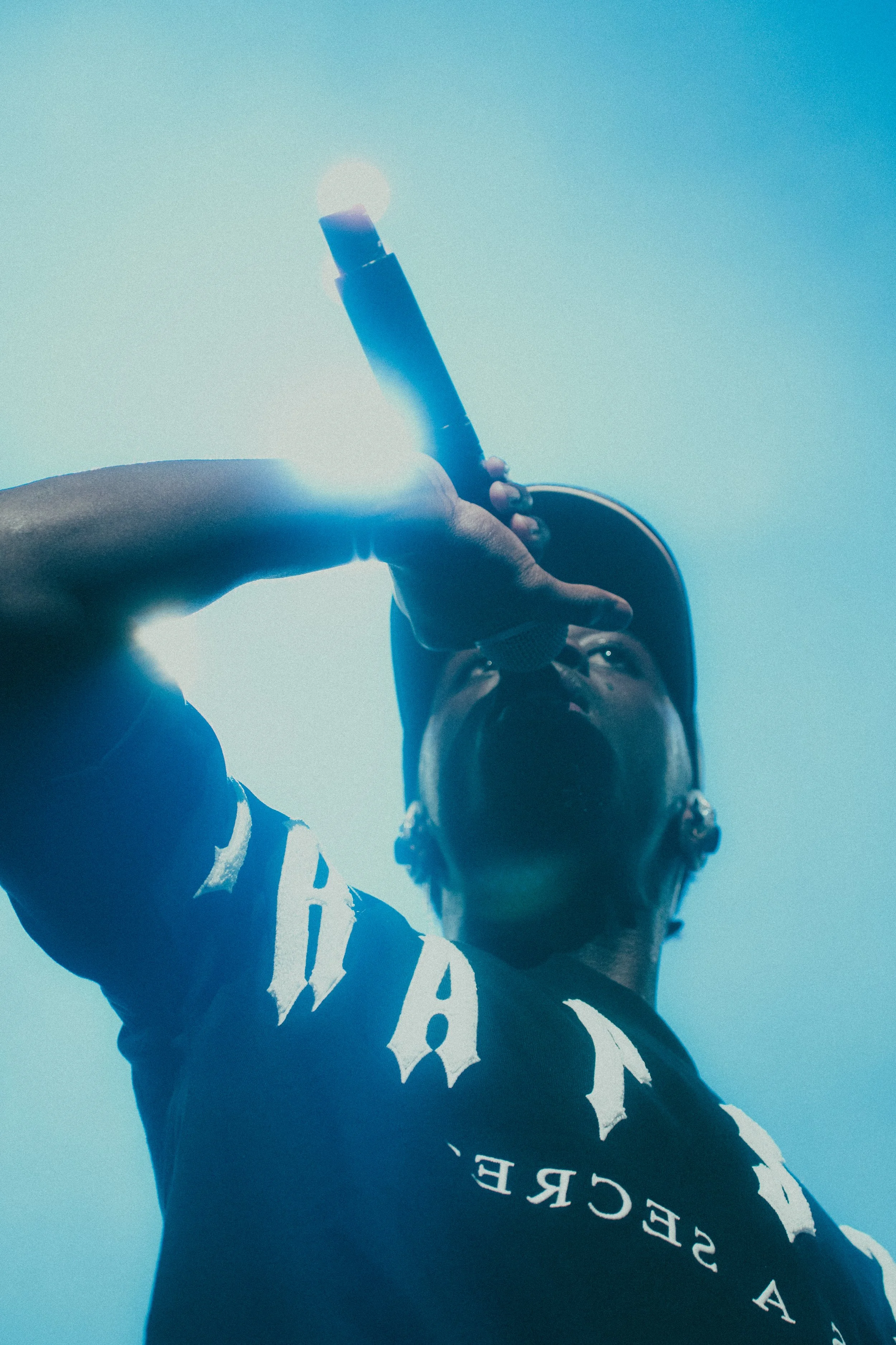 Une personne portant une casquette et un t-shirt noir tient un microphone ou un objet cylindrique près de la bouche, vue de dessous avec un ciel bleu en arrière-plan.