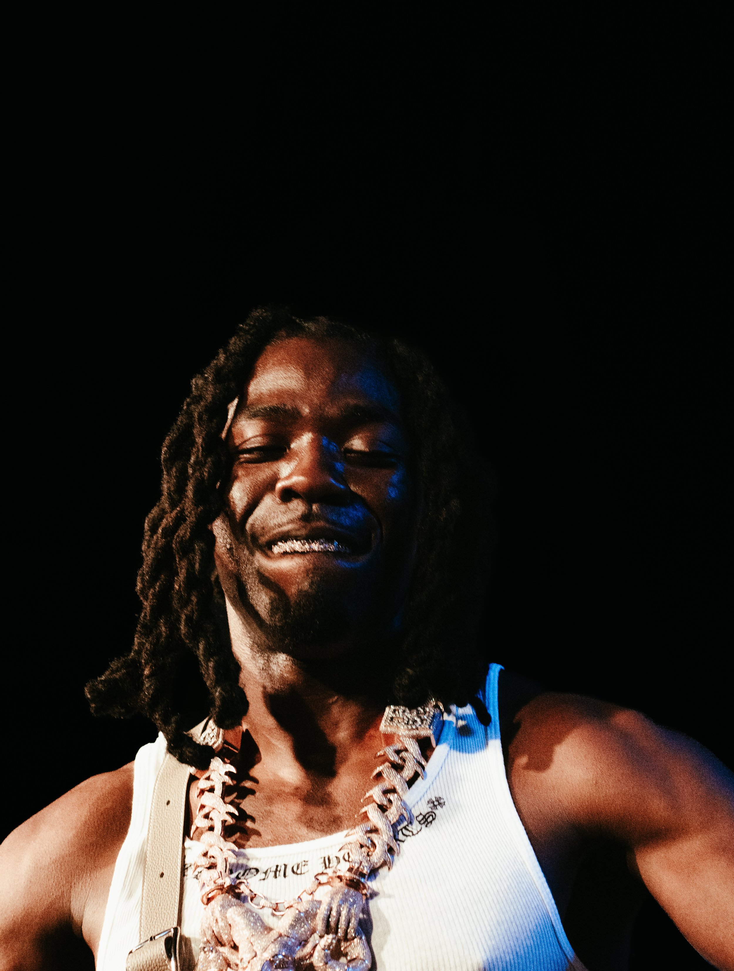 Un homme souriant portant un collier en coquillages, un t-shirt blanc, avec des dreadlocks, dans un environnement sombre.