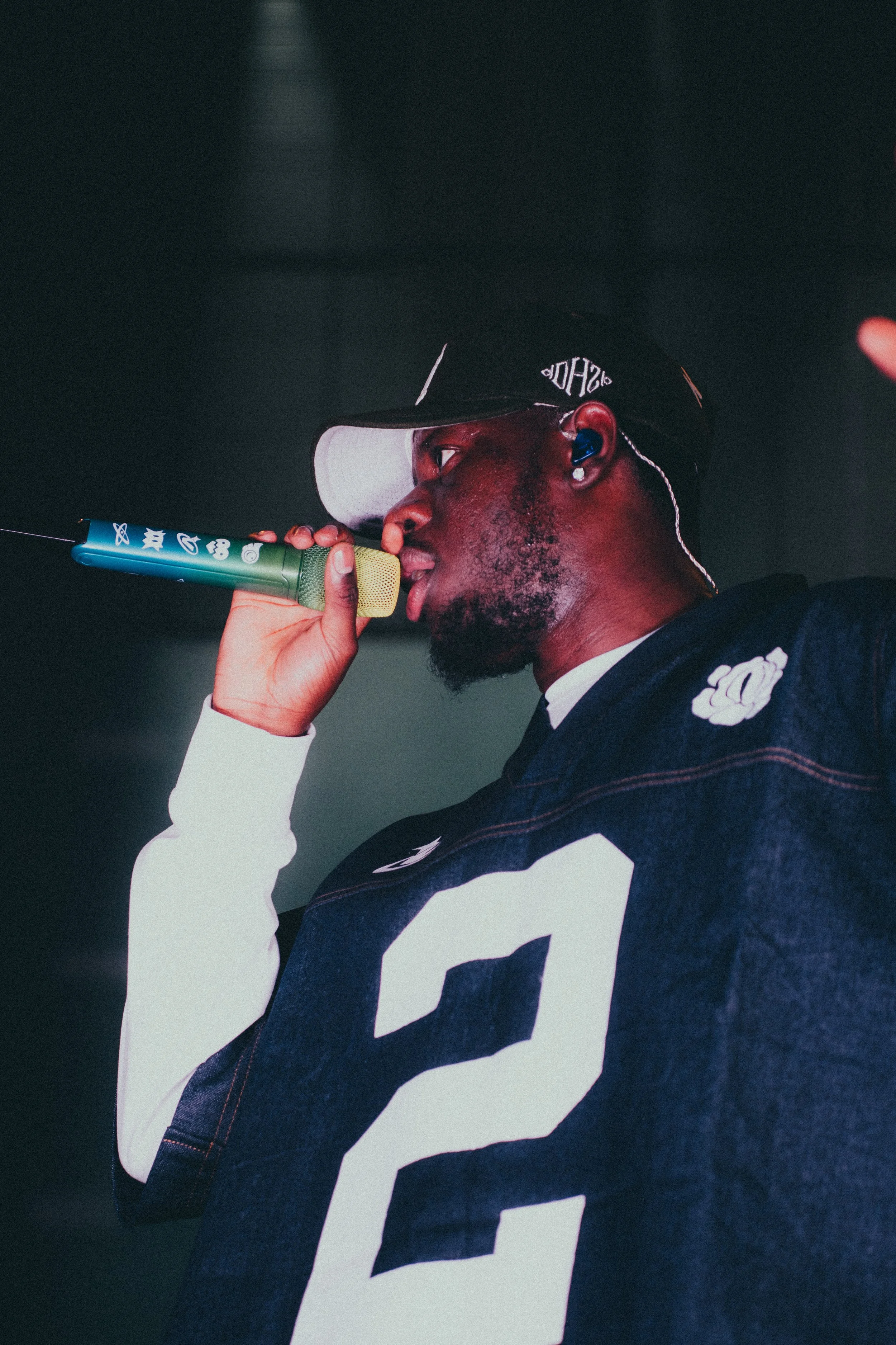Un homme noir portant une casquette noire, des écouteurs bleus, un maillot de sport avec le numéro 2, tient un microphone vert et bleu, et chante ou parle.