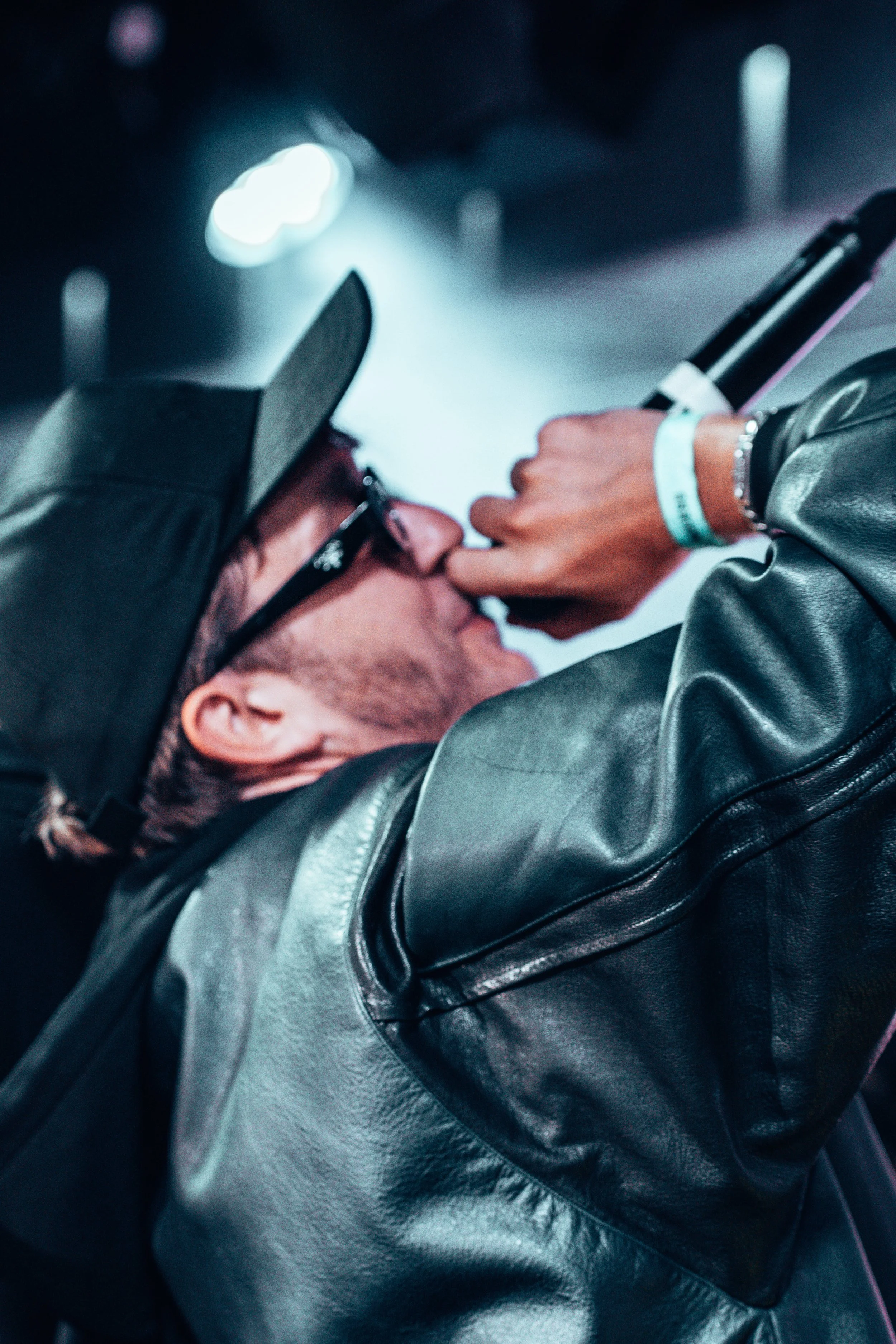 Un homme portant des lunettes, une casquette et une veste en cuir noir, tenant un microphone et chantant ou parlant en public, avec un éclairage de scène en fond.