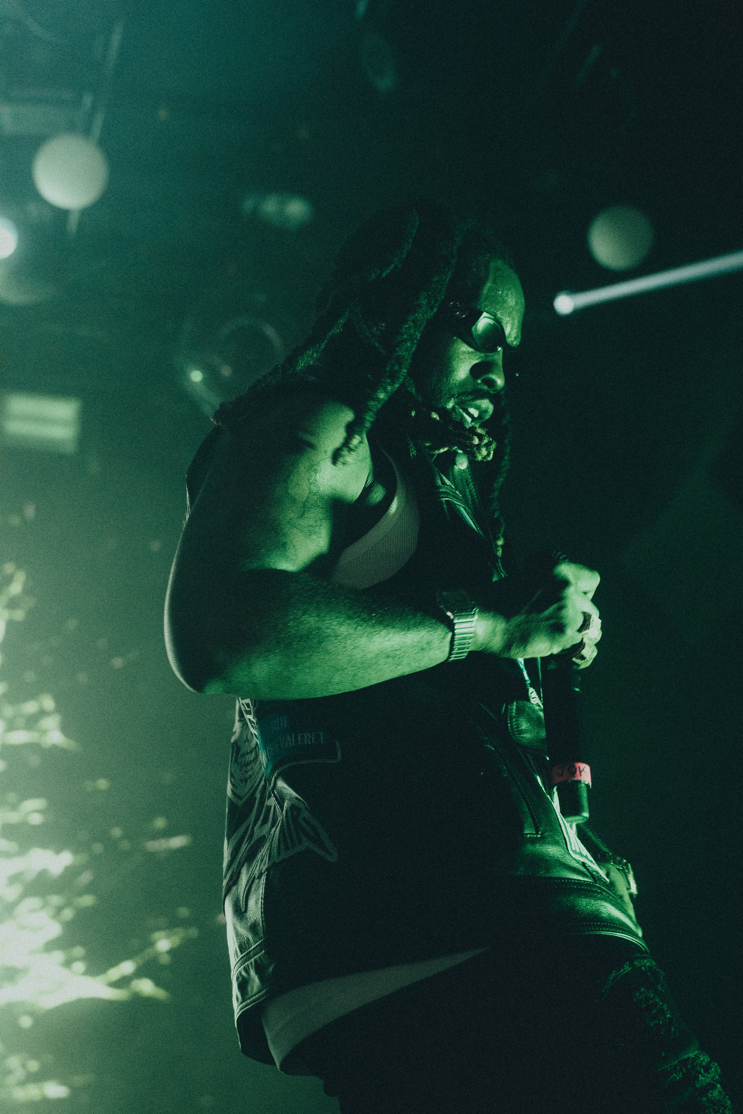 Un homme avec des dreadlocks portant des lunettes de soleil, tenant un micro sur scène, lumière verte dans l'ambiance d'un concert.