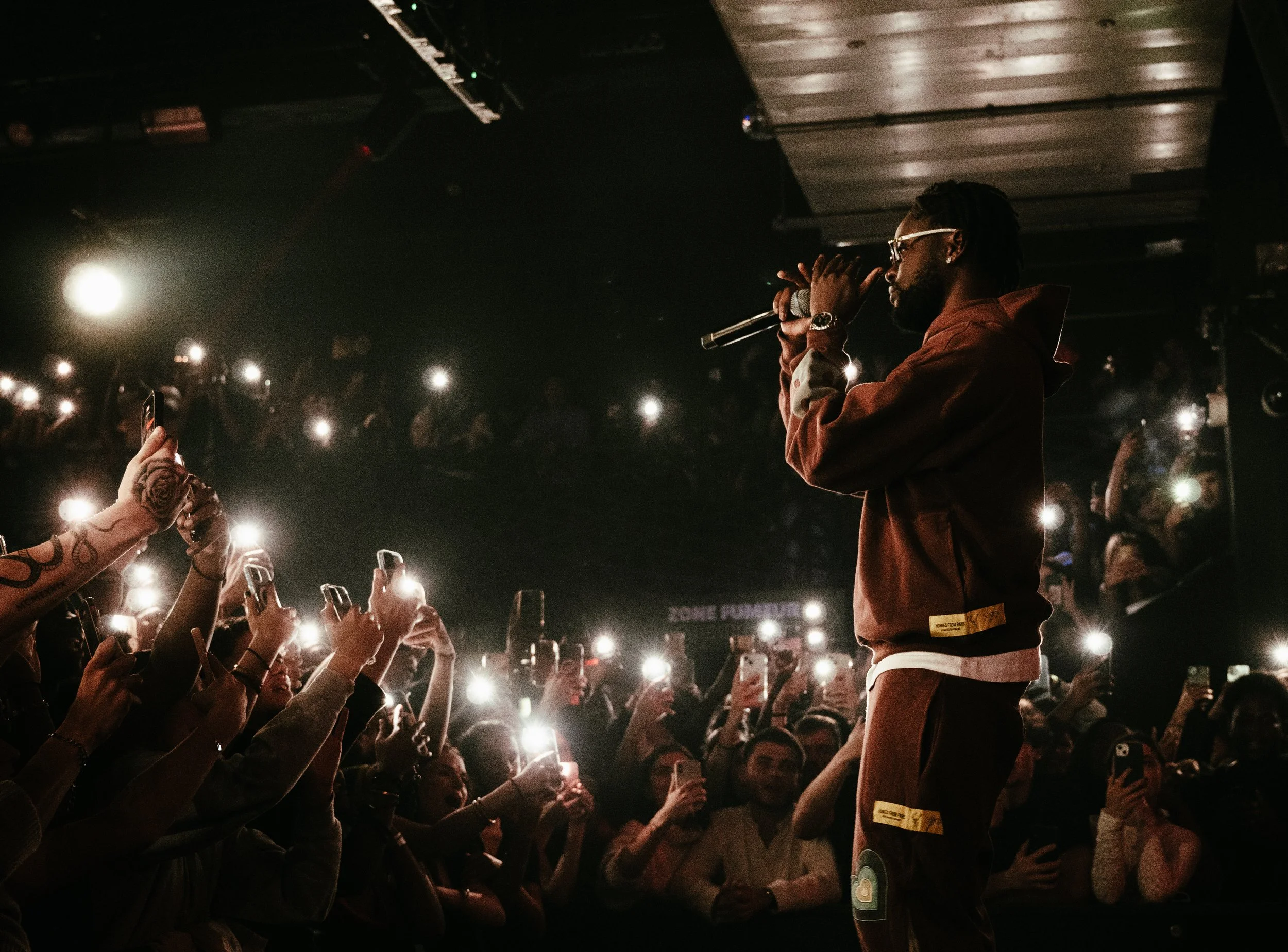 Un homme rappeur sur scène lors d'un concert, entouré d'une foule d'admirateurs qui utilisent leur téléphone et leur éclairage pour l'immortaliser.