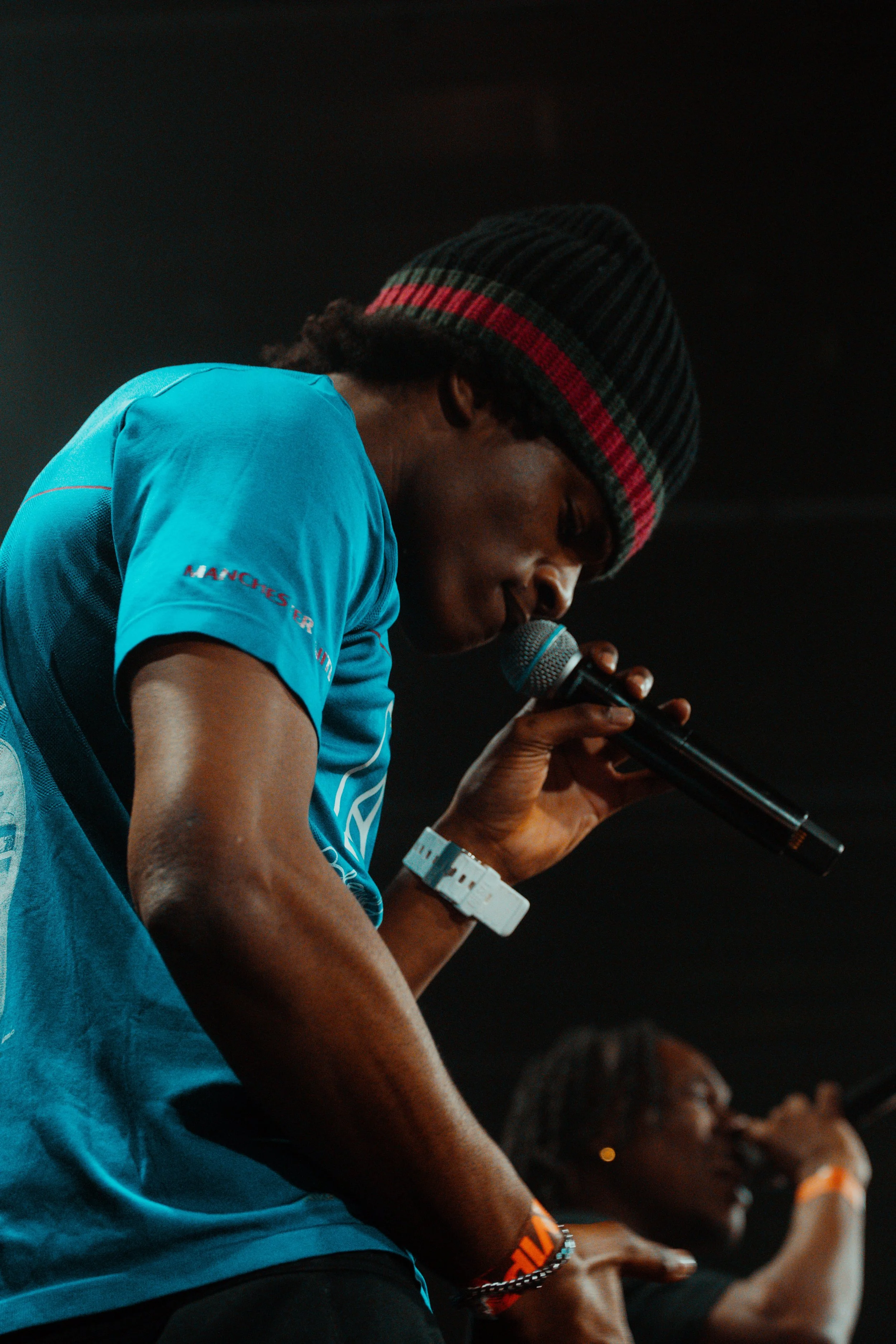 Un jeune homme avec une casquette noire à rayures rouges et vertes chante dans un microphone sur scène, avec une autre personne en arrière-plan.