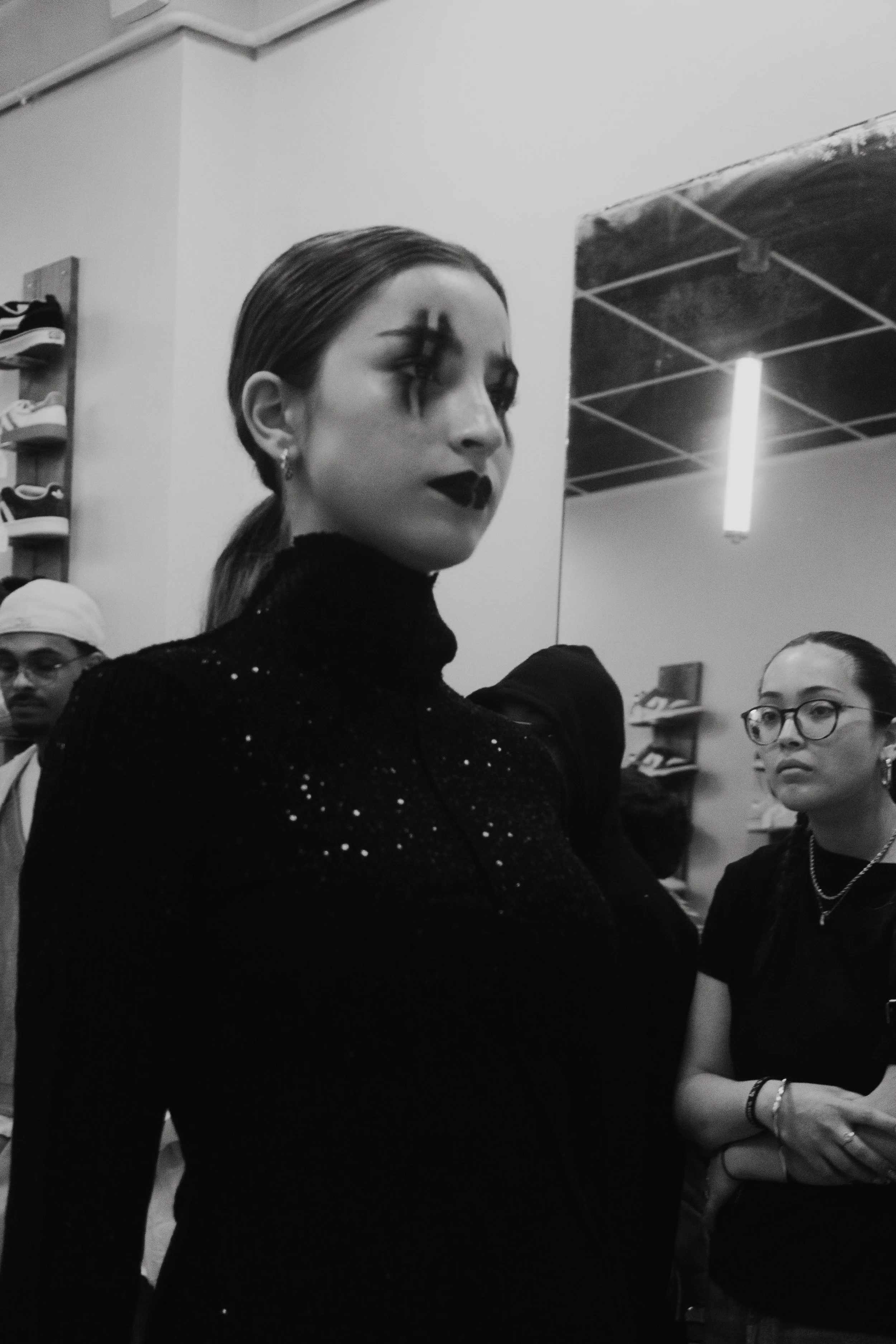 Une femme avec un maquillage sombre et des yeux maquillés de noir, portant un pull noir avec des étoiles scintillantes, en groupe avec d'autres personnes dans un magasin. L'image est en noir et blanc.