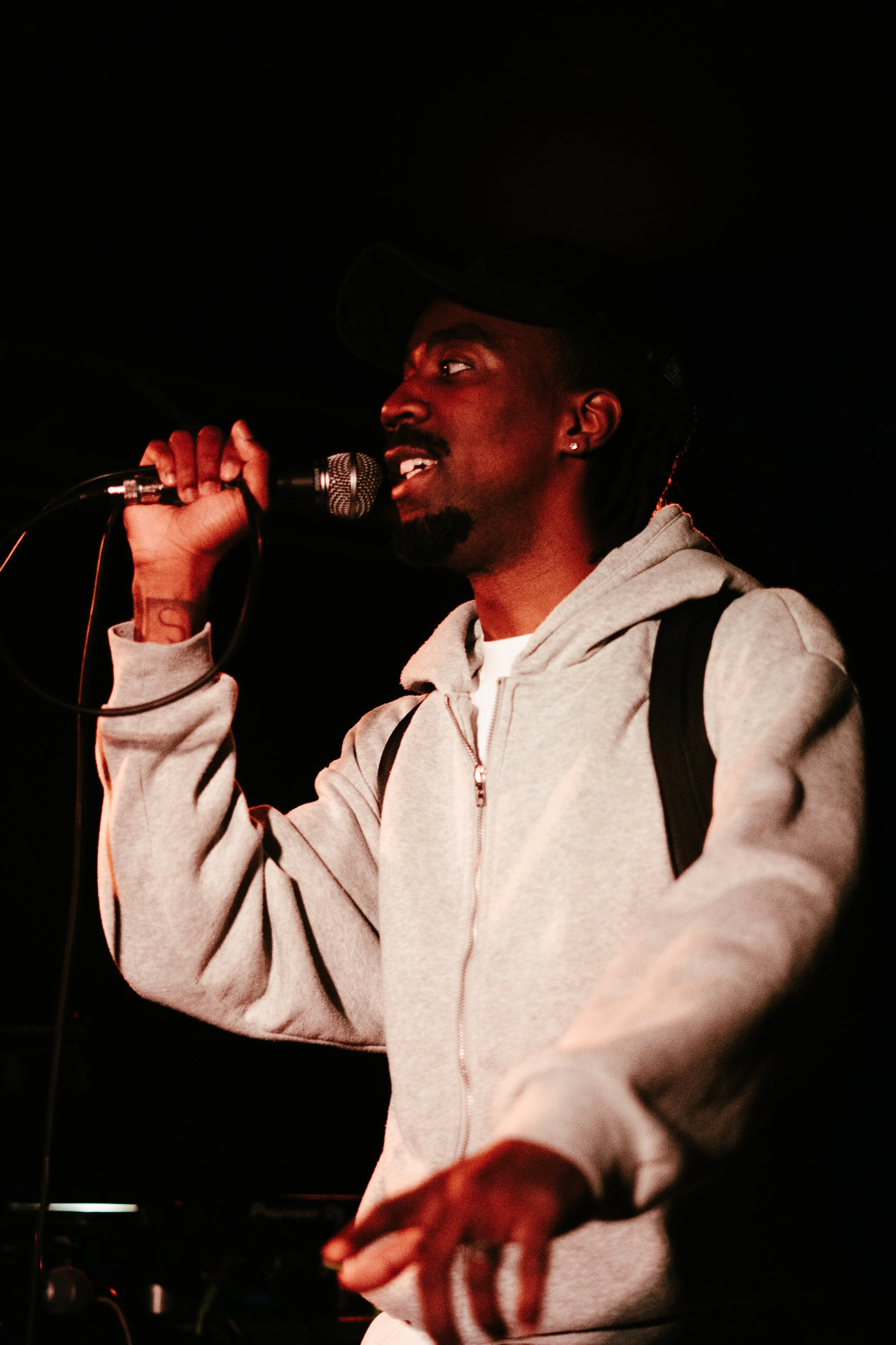 Un homme noir portant une casquette noire, une veste grise à capuche et un sac à dos, parlant ou chantant dans un microphone dans un environnement sombre.