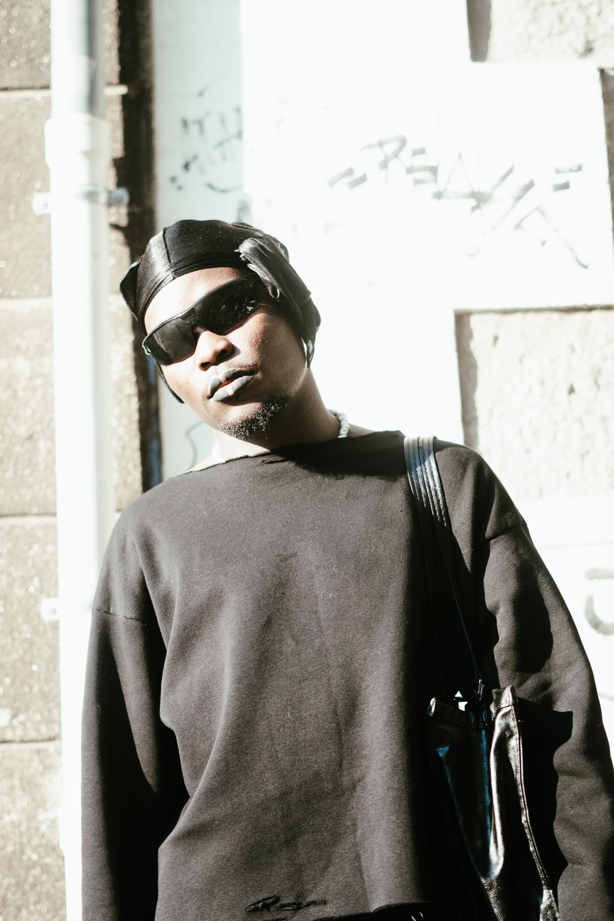 Un homme portant des lunettes de soleil noires, un bandeau noir, un sweat noir avec un sac noir en bandoulière, se tenant devant un mur en pierre avec des graffitis.