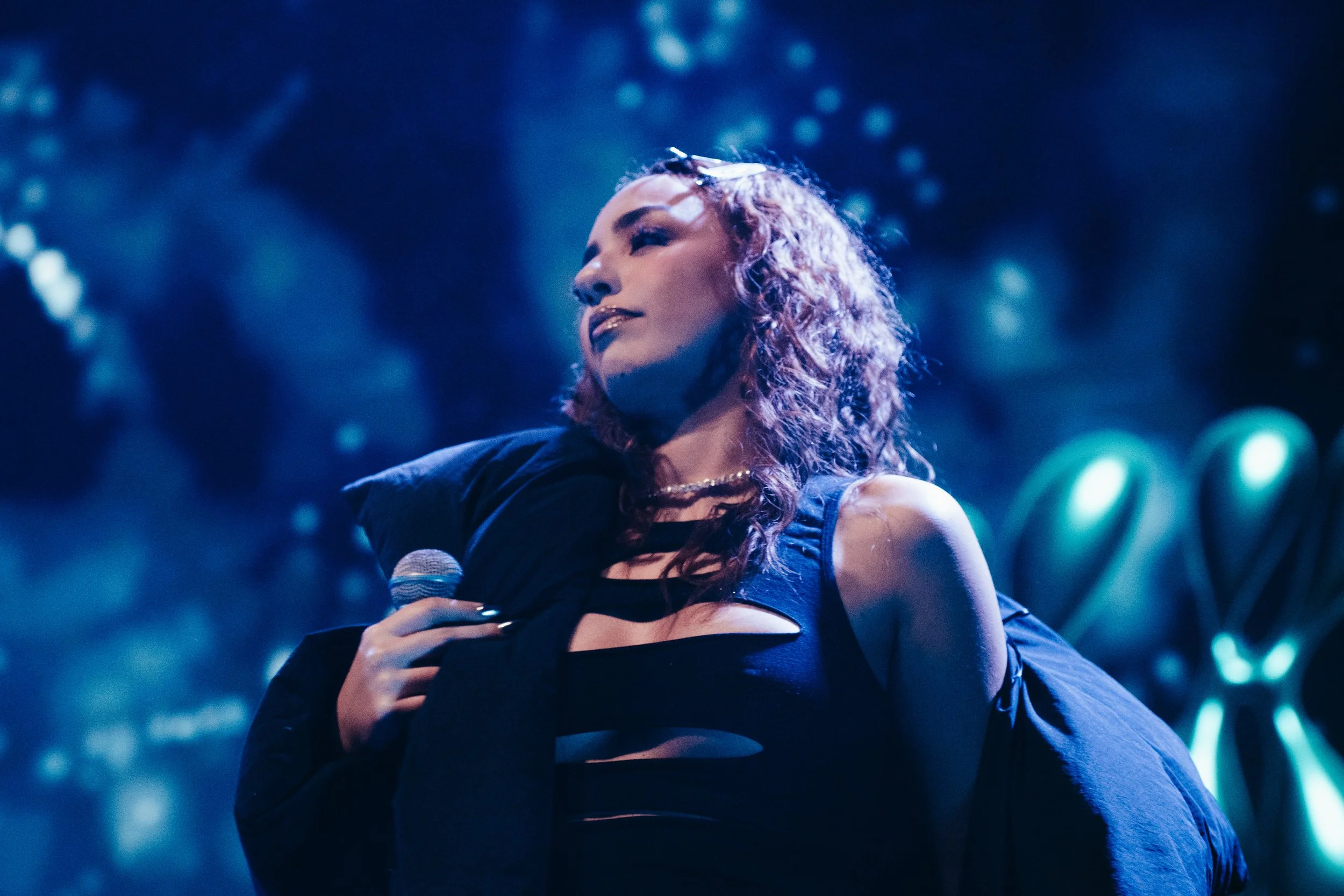 Une femme avec des cheveux bouclés rouges, tenant un microphone, sur scène avec un éclairage bleu et nuit artificielle