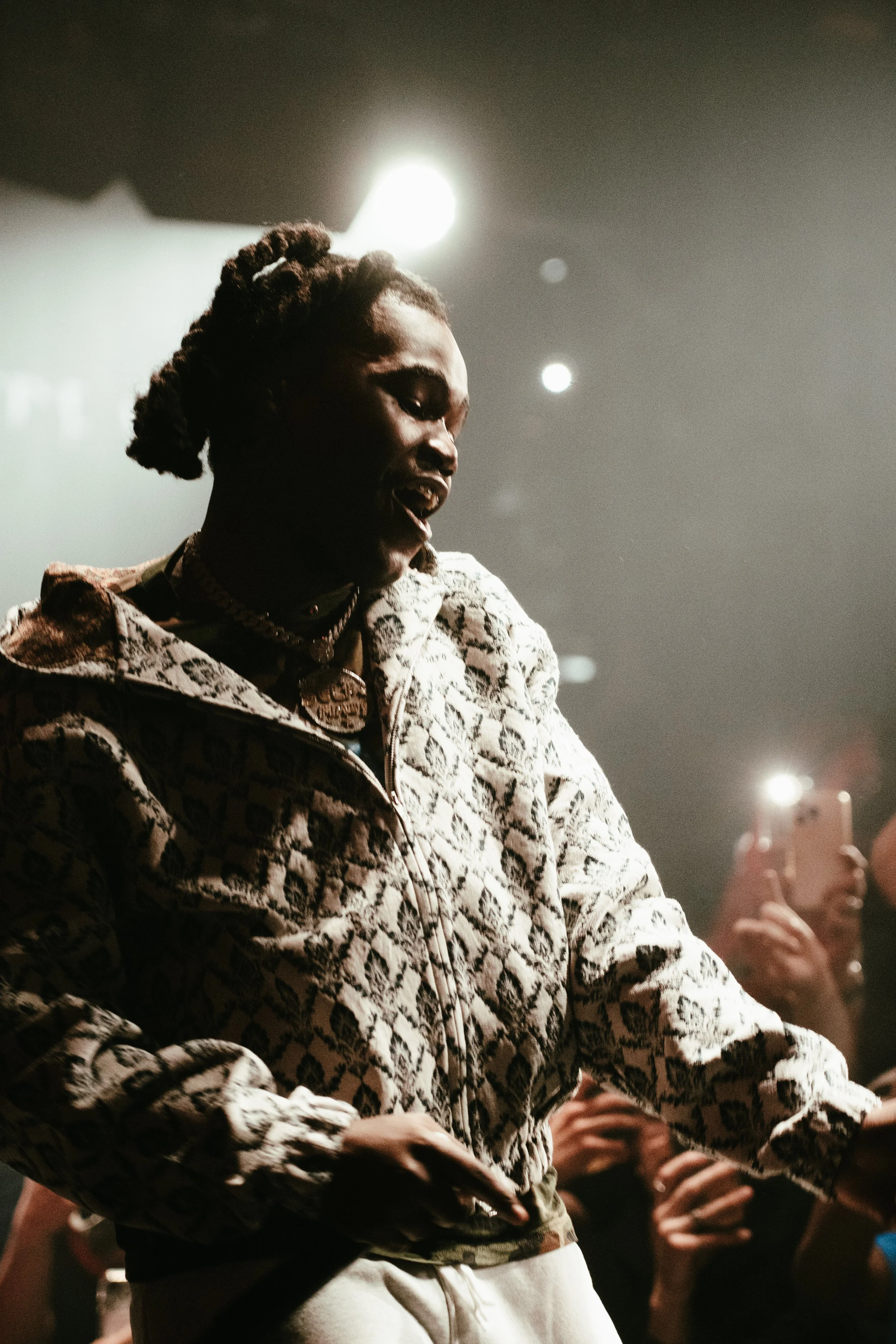 Femme noire dans un éclairage sombre, portant une veste à motif, en train de danser ou de se déhancher lors d'une performance ou d'une soirée, avec des personnes au fond utilisant des téléphones.