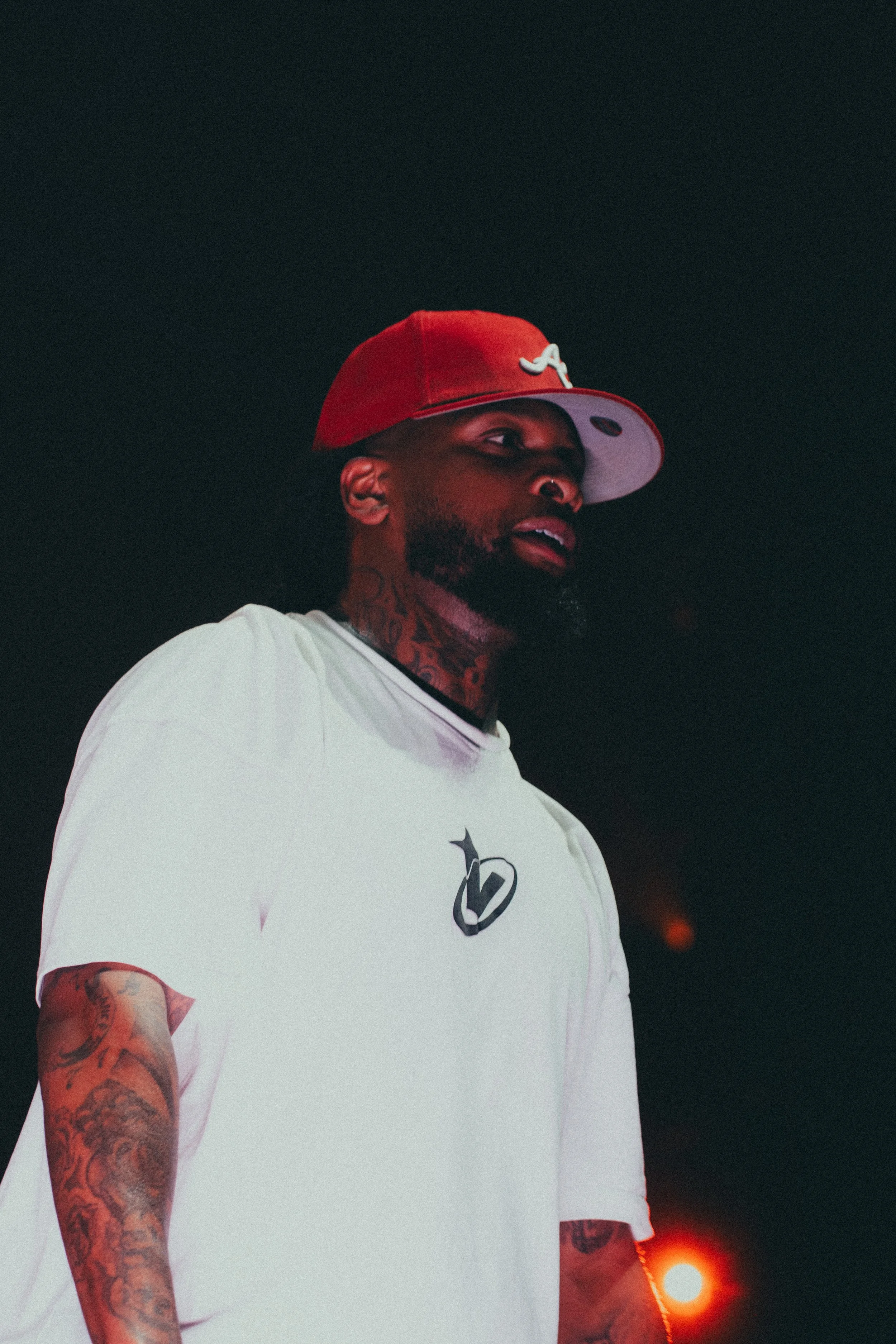 Un homme portant un t-shirt blanc et une casquette rouge en train de regarder vers la droite, avec un fond noir et une lumière orange floue en arrière-plan.