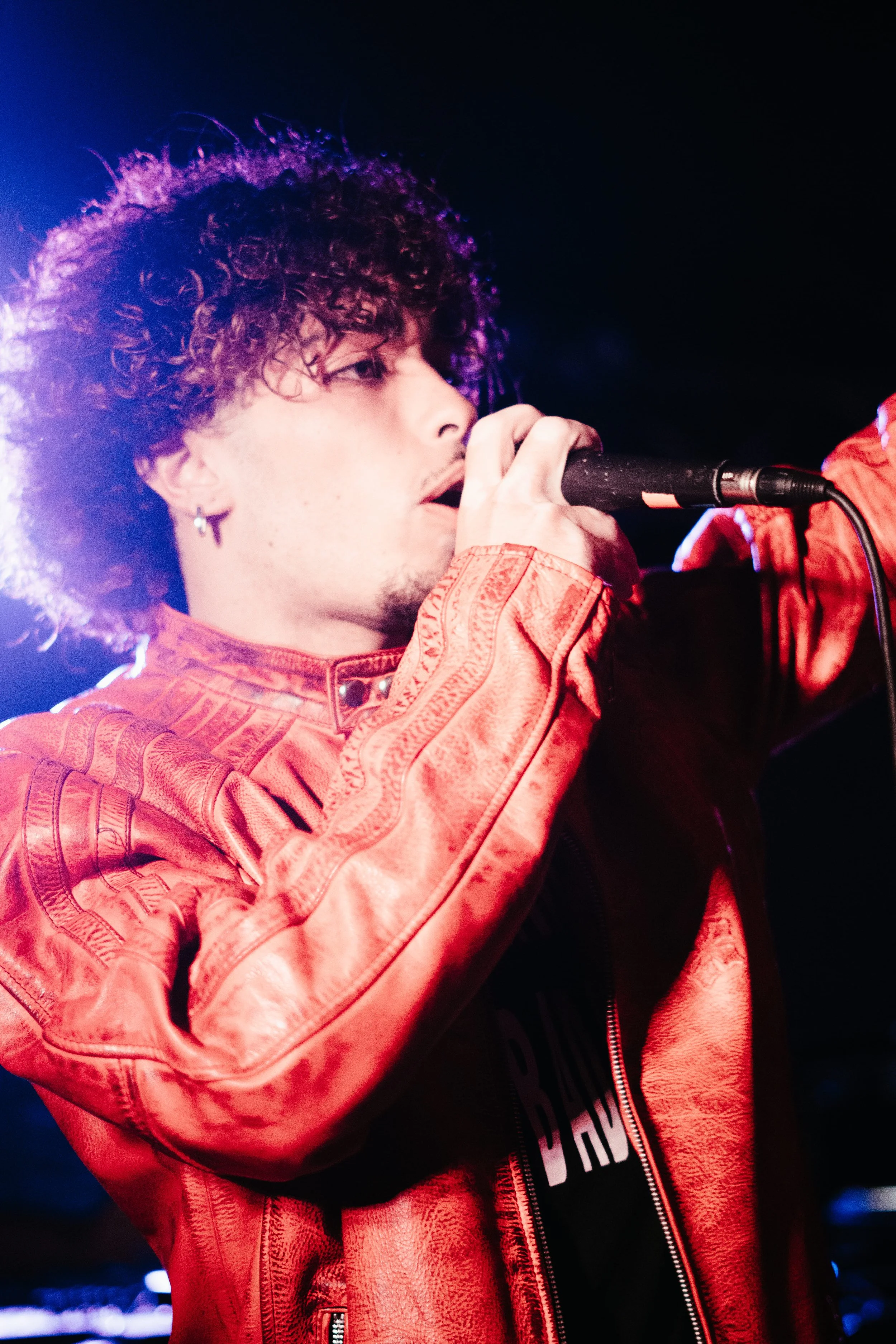 Un jeune homme avec des cheveux bouclés, portant une veste en cuir rouge, chante dans un micro avec un éclairage coloré dans le fond.