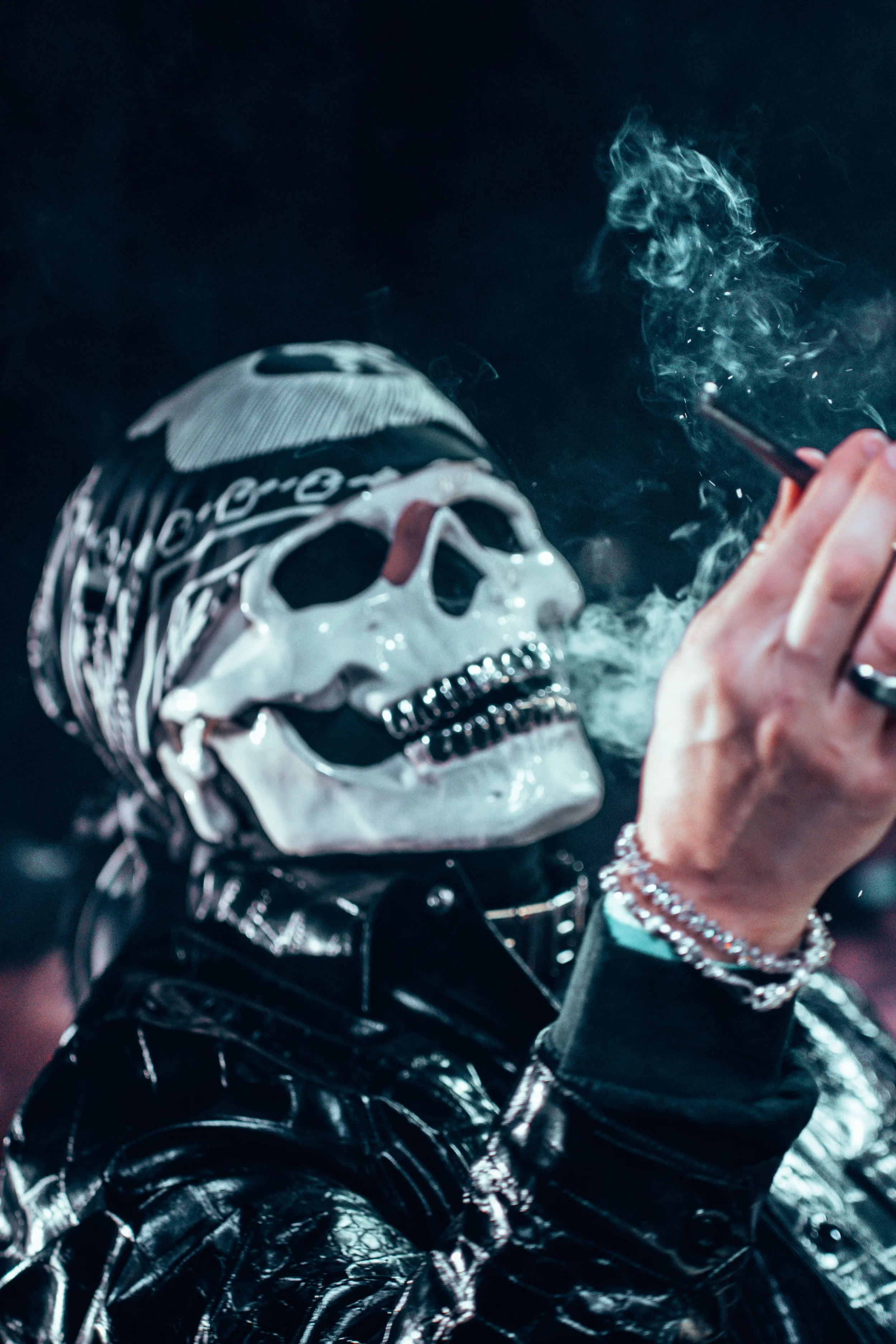 Personne portant un masque de crâne et un bandana, vêtu de cuir noir, en train de fumer une cigarette dans un environnement sombre.