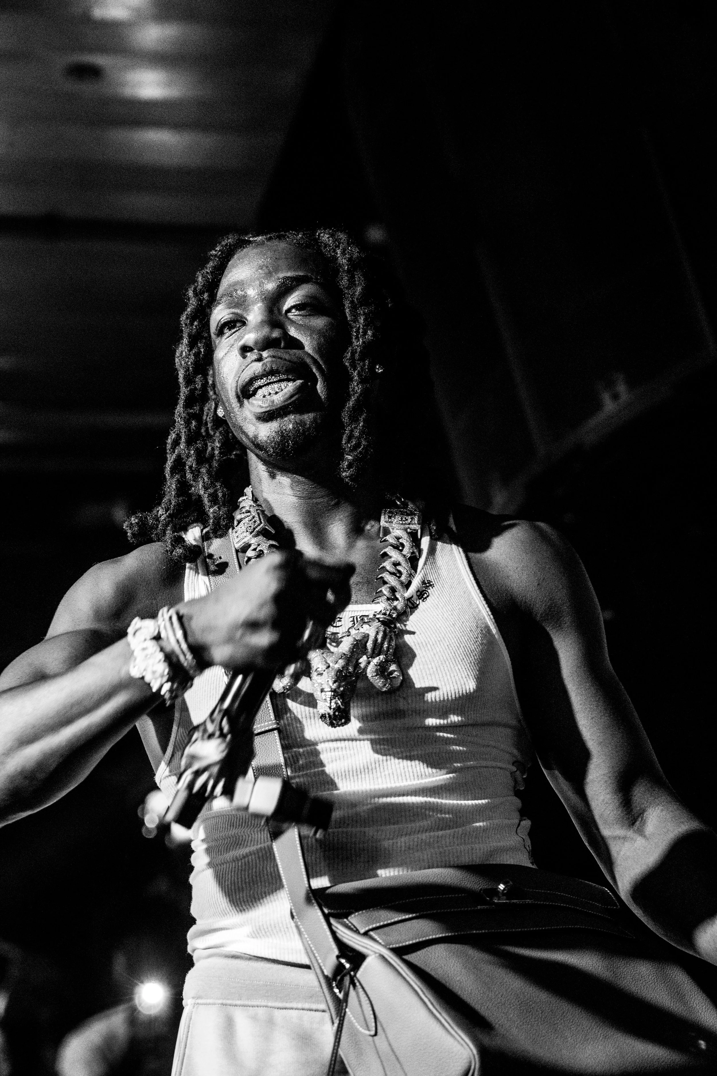 Un homme noir avec dreadlocks portant un t-shirt sans manches, des bijoux, et tenant un micro, en train de performer dans un environnement sombre.