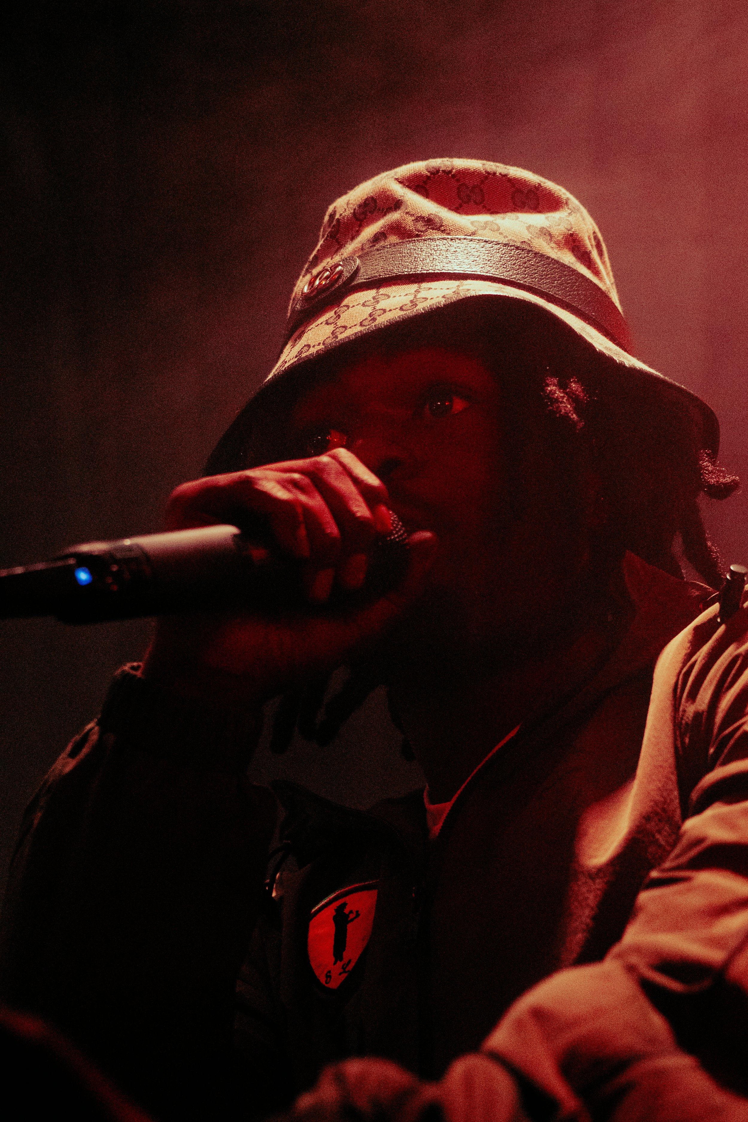 Un homme avec un chapeau et une veste, tenant un microphone, dans une lumière rougeâtre.