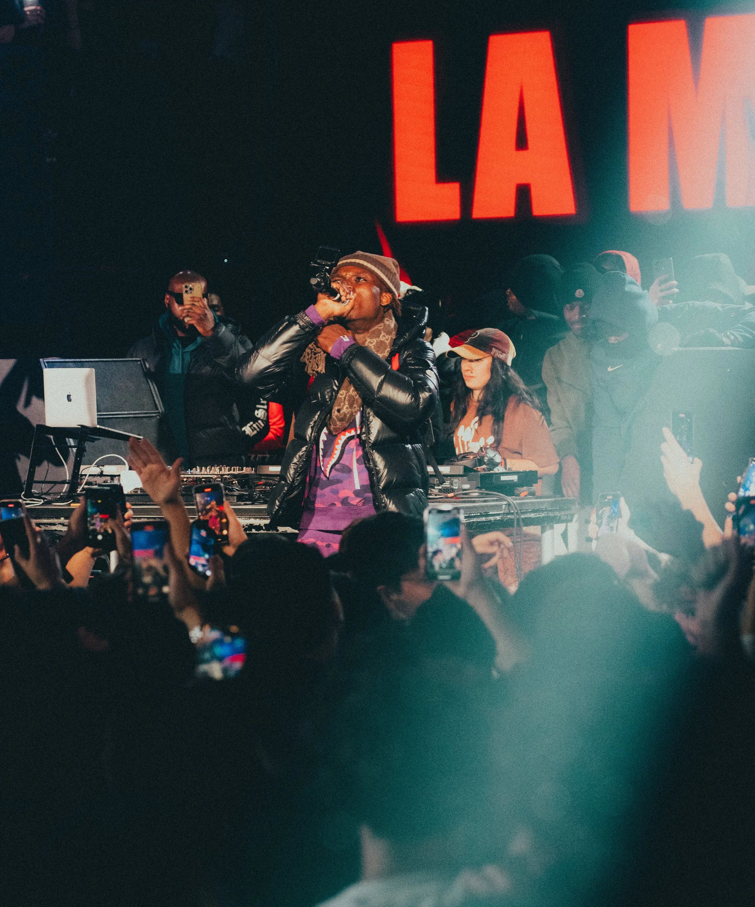 Un artiste hip-hop performe sur scène lors d'un concert avec un groupe, entouré de nombreux spectateurs qui prennent des photos et vidéos avec leurs téléphones.