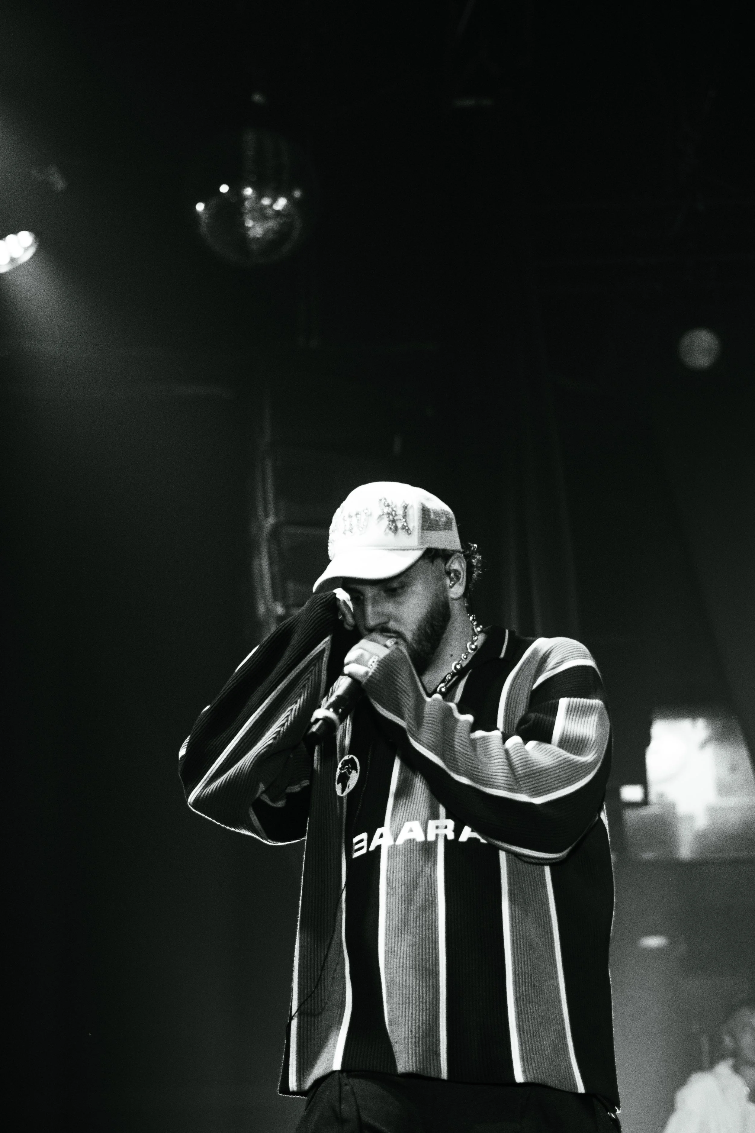 Un homme avec une casquette blanche chantant ou parlant dans un microphone lors d'une performance, en noir et blanc.