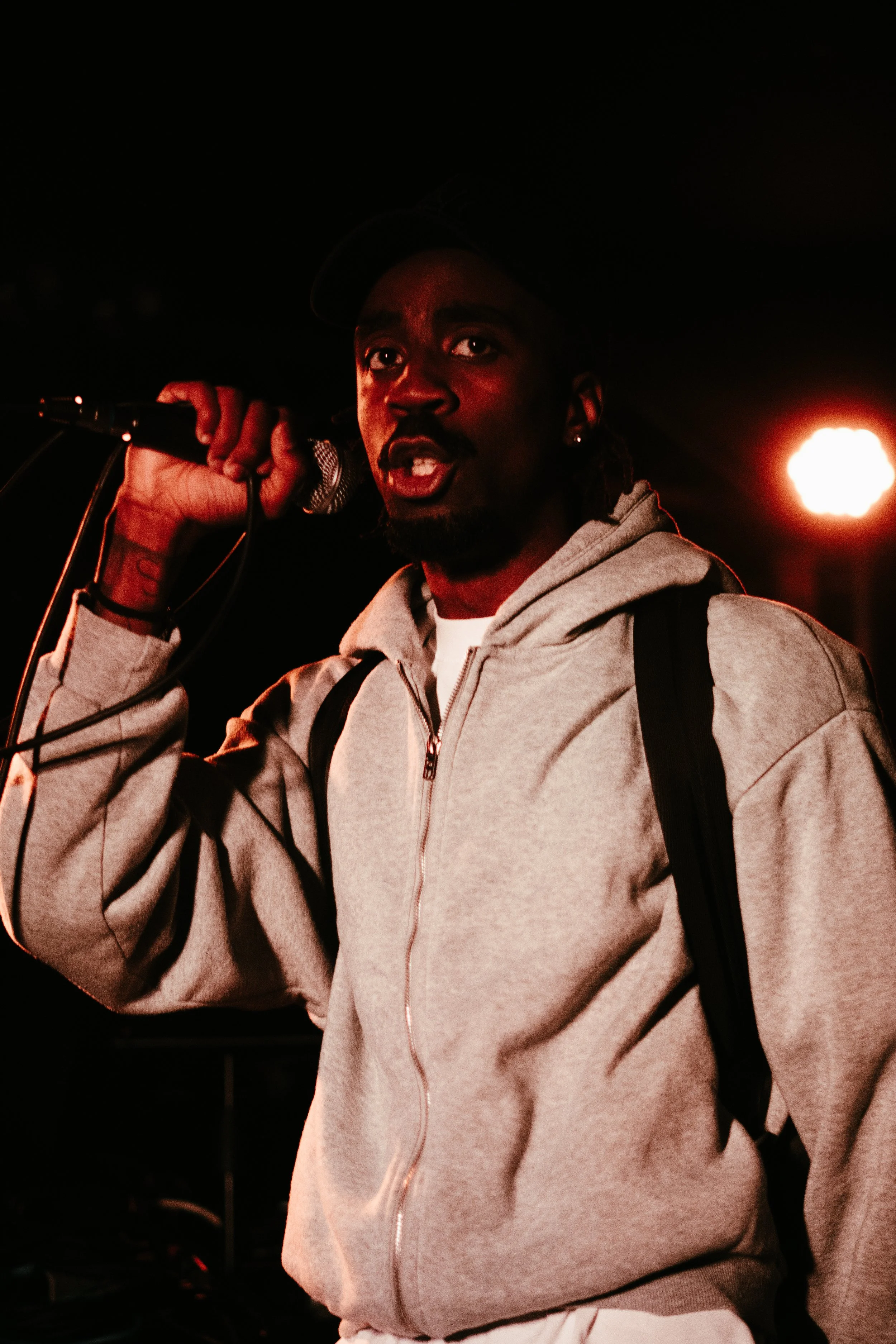 Un homme chante dans un microphone, portant un sweat à capuche gris et un sac à dos, avec un éclairage rouge en arrière-plan.
