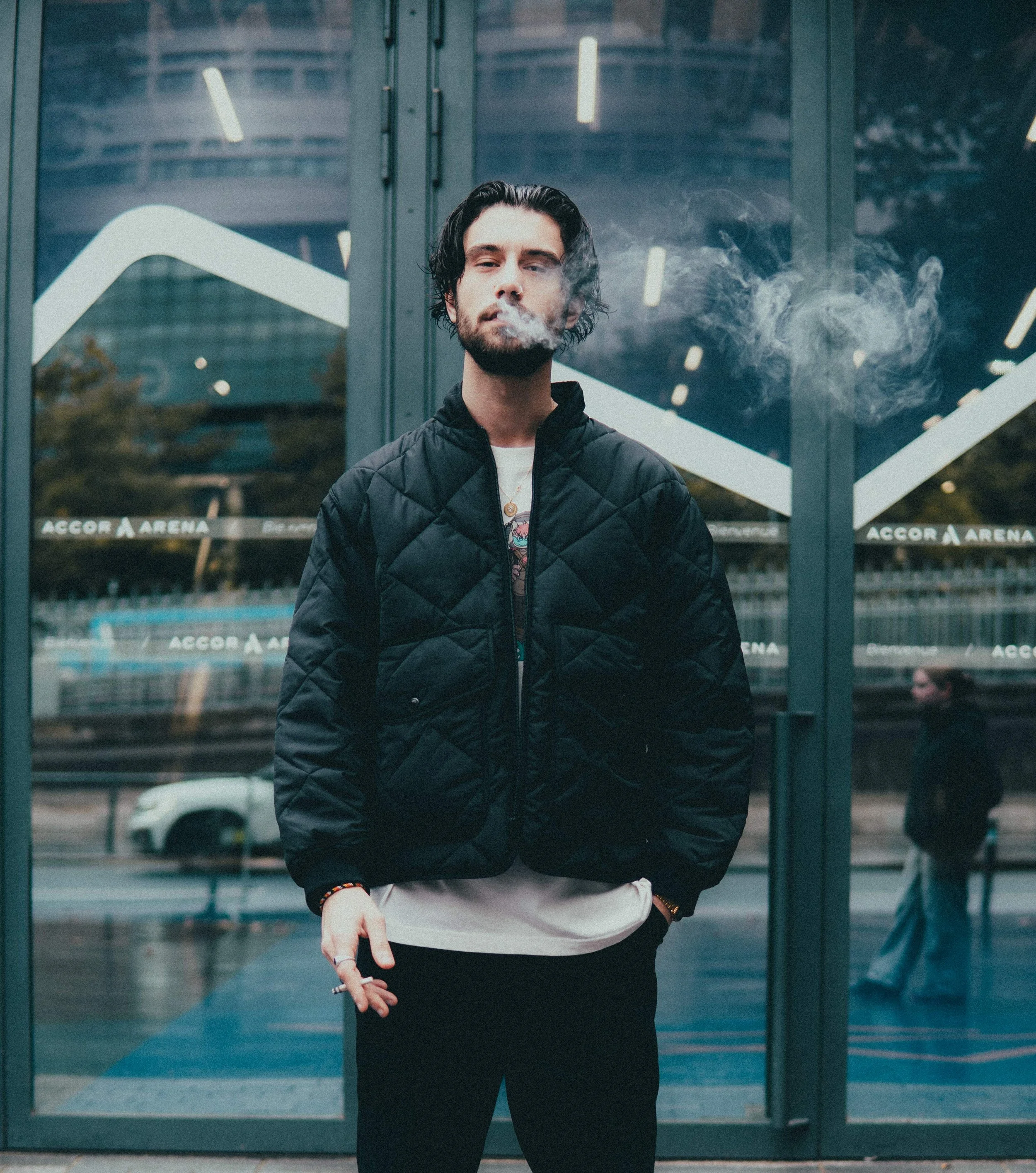 Un homme moustachu en veste noire et t-shirt blanc devant une porte en verre, fumant une cigarette, avec un bâtiment en arrière-plan.