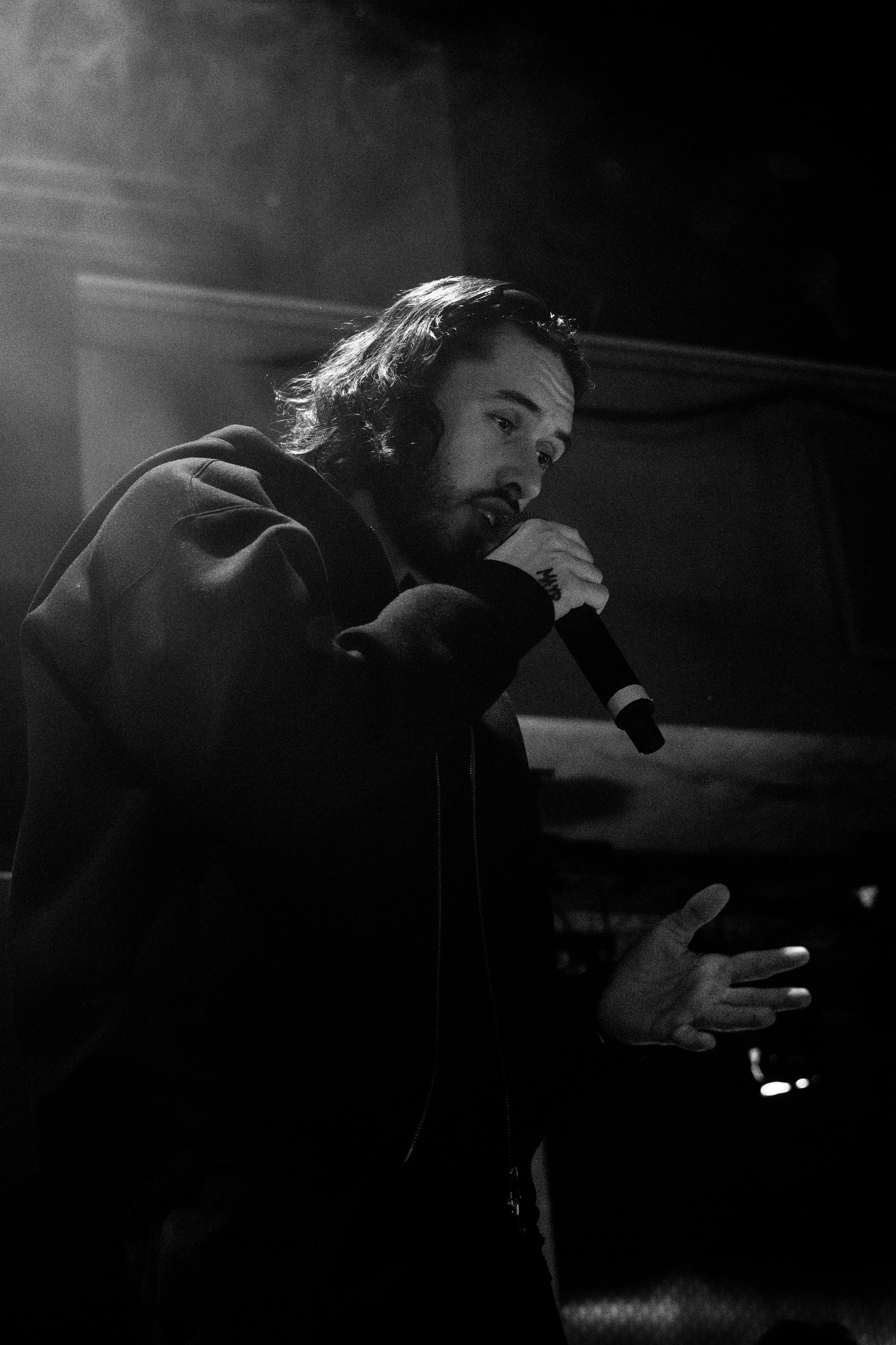Un homme avec de longs cheveux et une barbe chante dans un micro lors d'une performance, en noir et blanc.