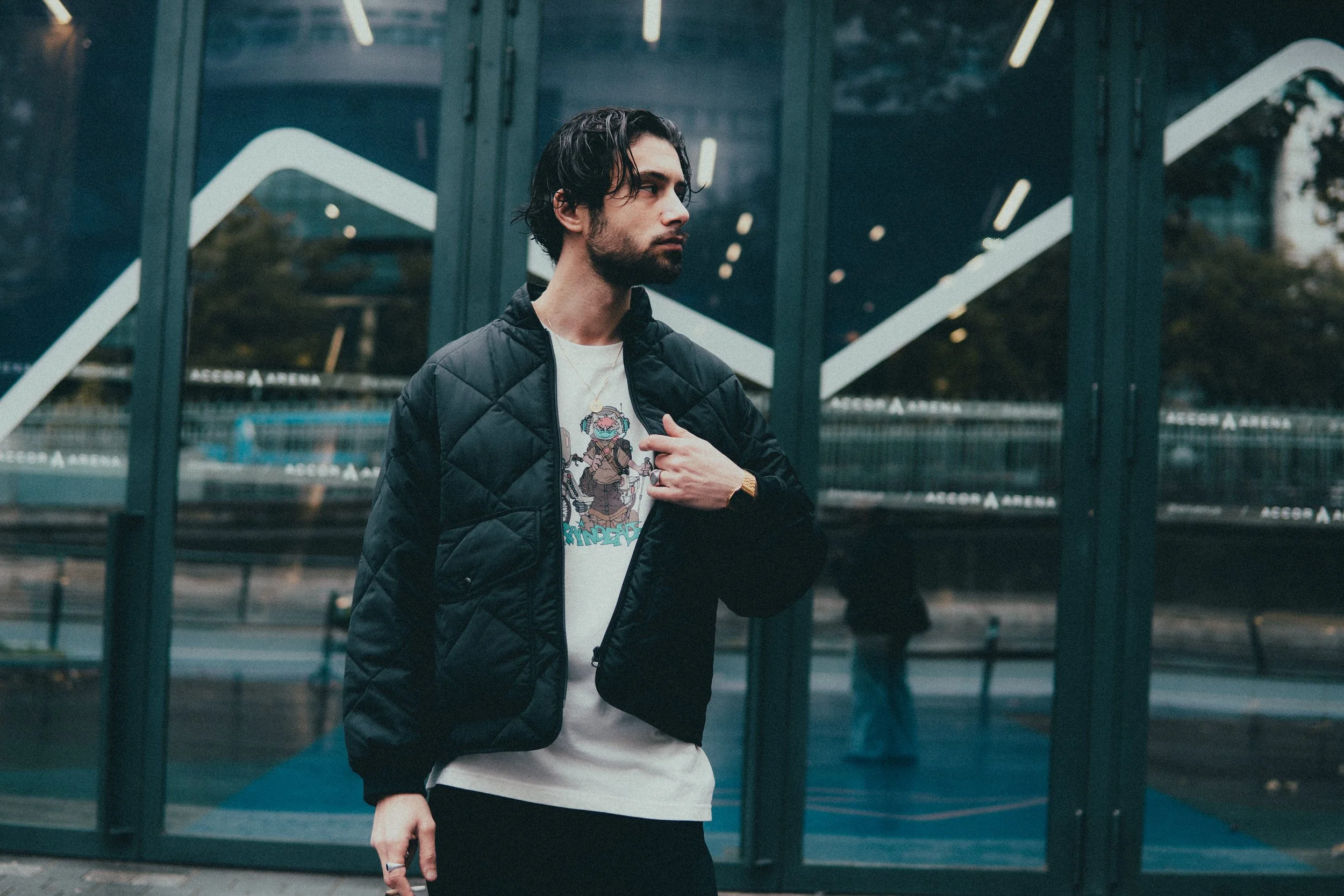Jeune homme avec une veste noire, t-shirt graphique, dans une zone urbaine devant une structure vitrée.