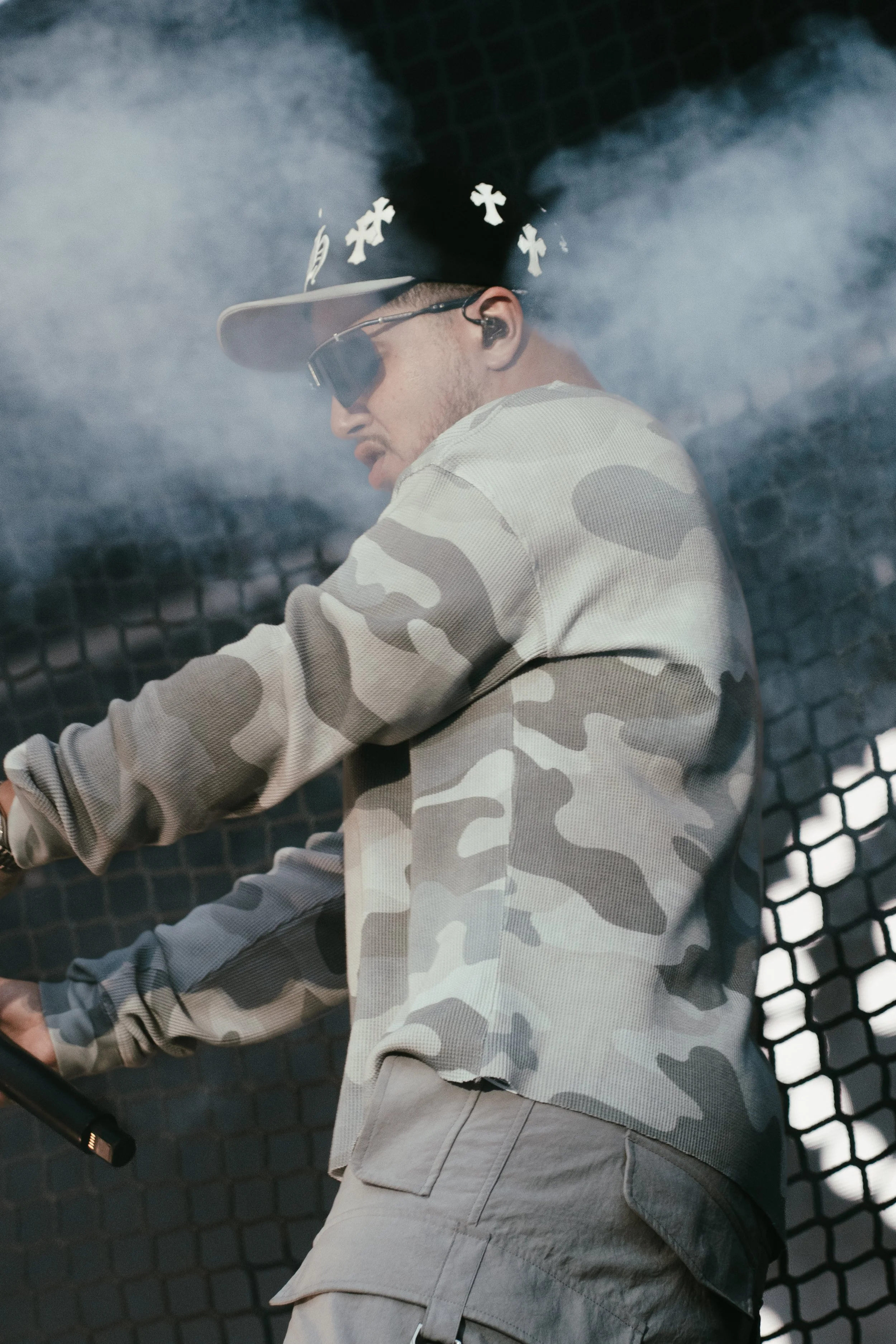 Un jeune homme portant un chapeau de style cowboy, des lunettes de soleil noires, un micro-cravate et une veste à motif camouflage gris, en train de jouer d'un instrument de musique devant un fond de grille noir avec de la fumée.