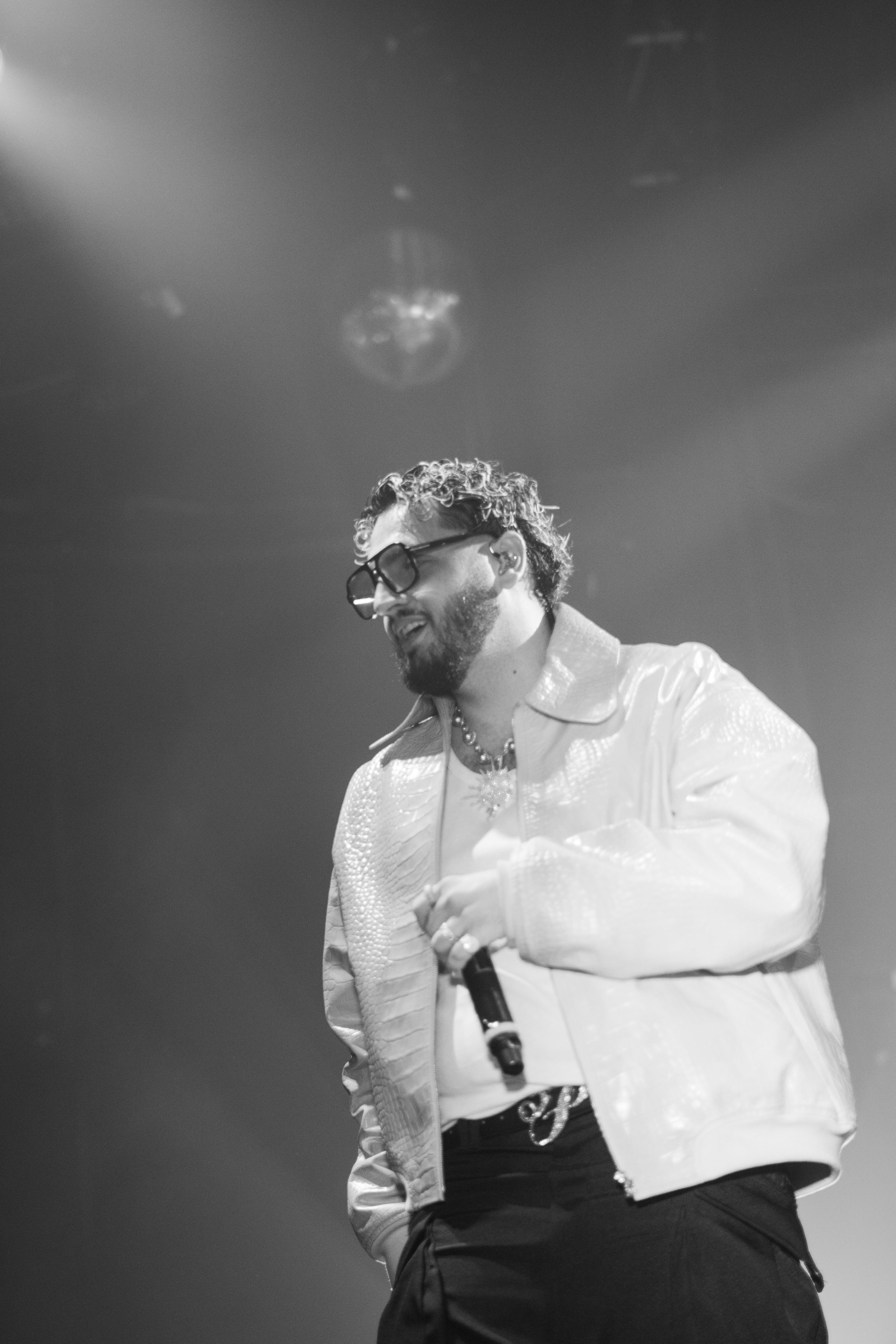 Un homme portant des lunettes noires, une veste blanche et un collier, tenant un microphone lors d'une performance sur scène.