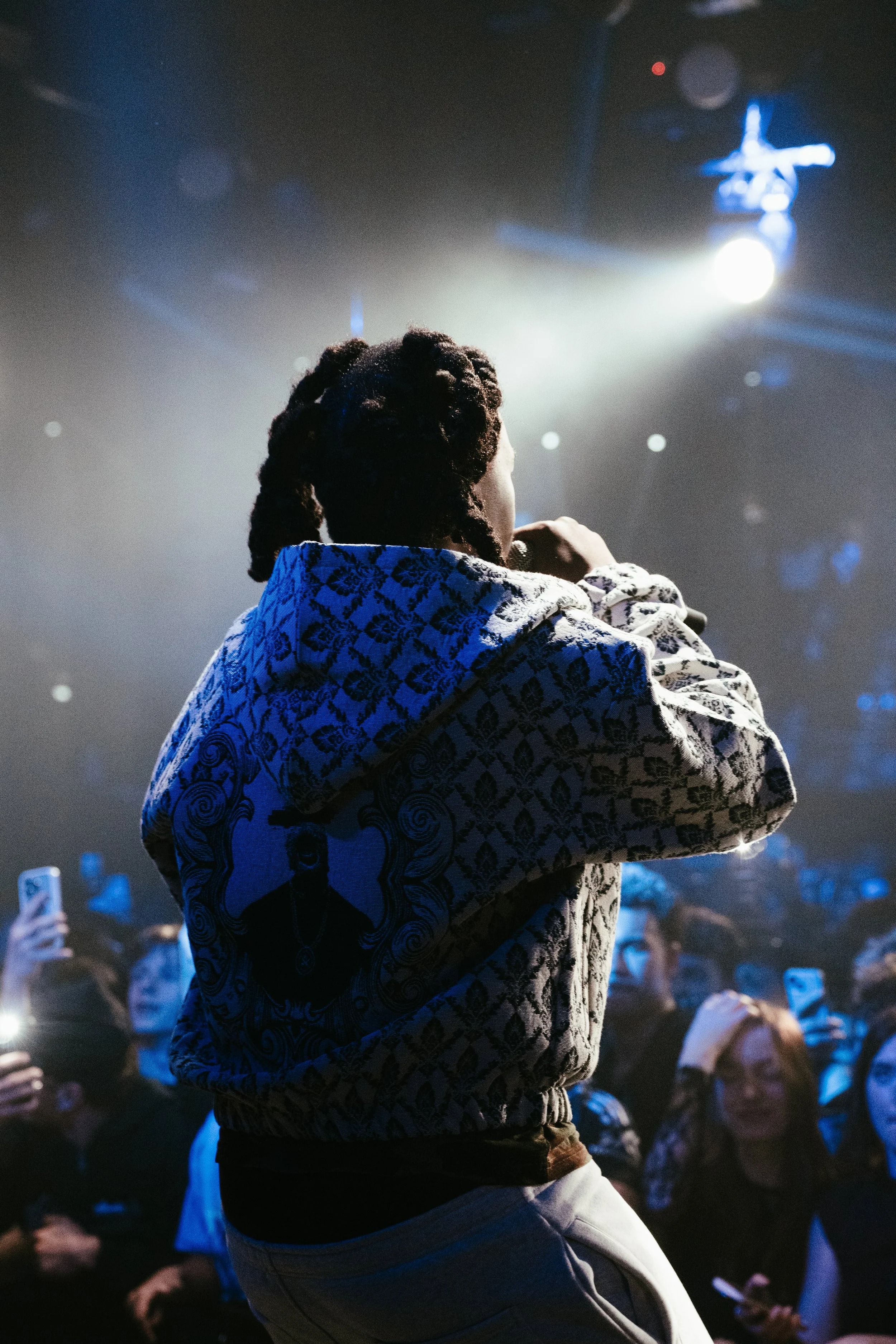 Une personne vue de dos, tenant un microphone, sur scène lors d'une performance ou d'un concert, avec un public devant elle et un éclairage de scène intense.
