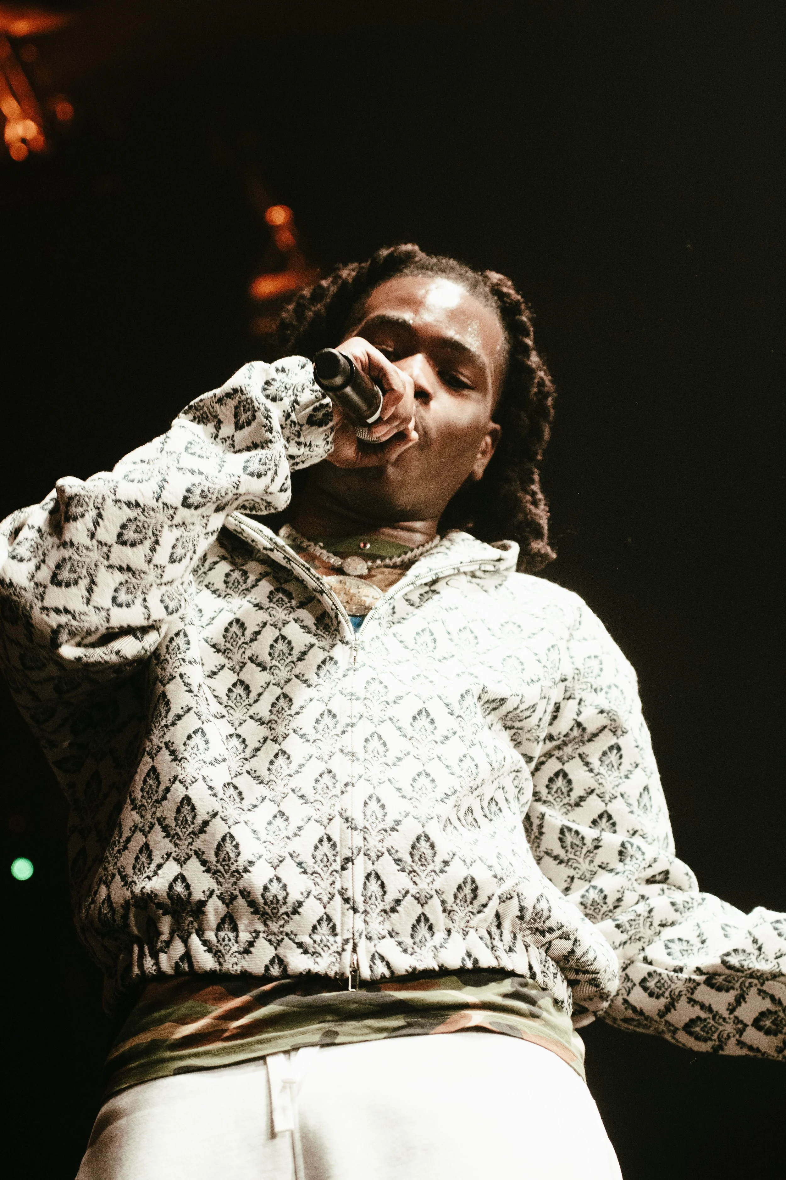 Jeune homme chantant en micro sur scène, portant un sweat blanc à motif noir, avec des bijoux, dans un éclairage de concert 