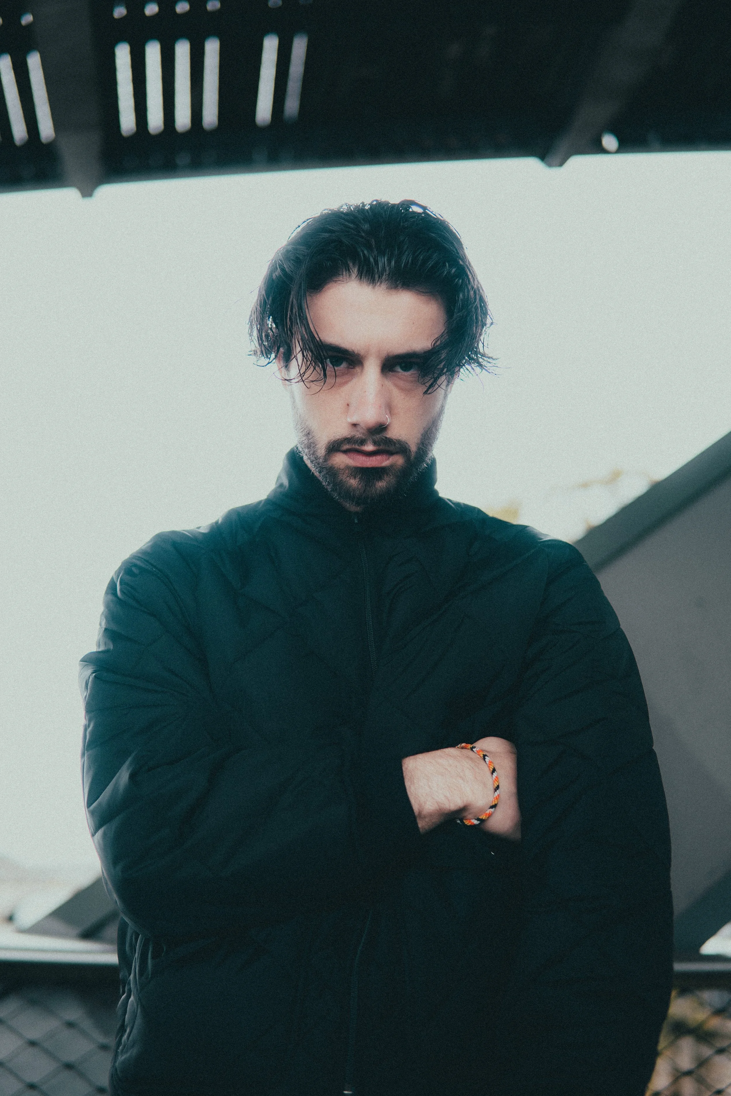 Un jeune homme avec des cheveux noirs, portant une veste noire, avec les bras croisés et un bracelet coloré au poignet, se tenant devant une structure en métal et un ciel clair.